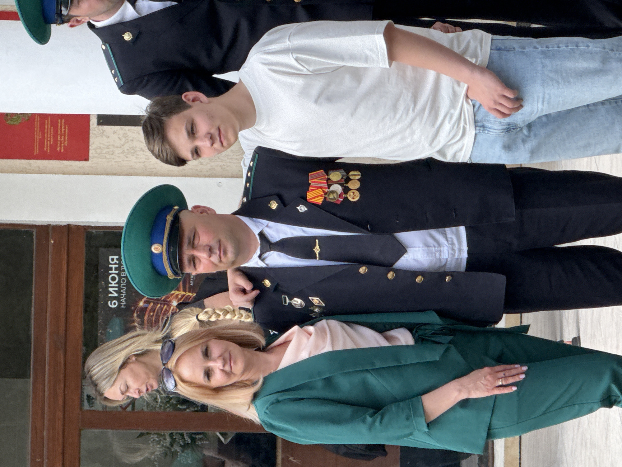 The image shows four people. A woman with blonde hair and sunglasses stands in the foreground, wearing a green blazer over a light pink top. Next to her is a man in a green military hat, wearing a black military jacket adorned with medals. Behind him is a younger man wearing a white T-shirt and light blue jeans. In the background, a man wearing sunglasses can be partially seen. They are standing in front of a building with a sign indicating "6 ИЮНЯ." The location is in an urban setting, likely in the city of Unknown, Unknown.