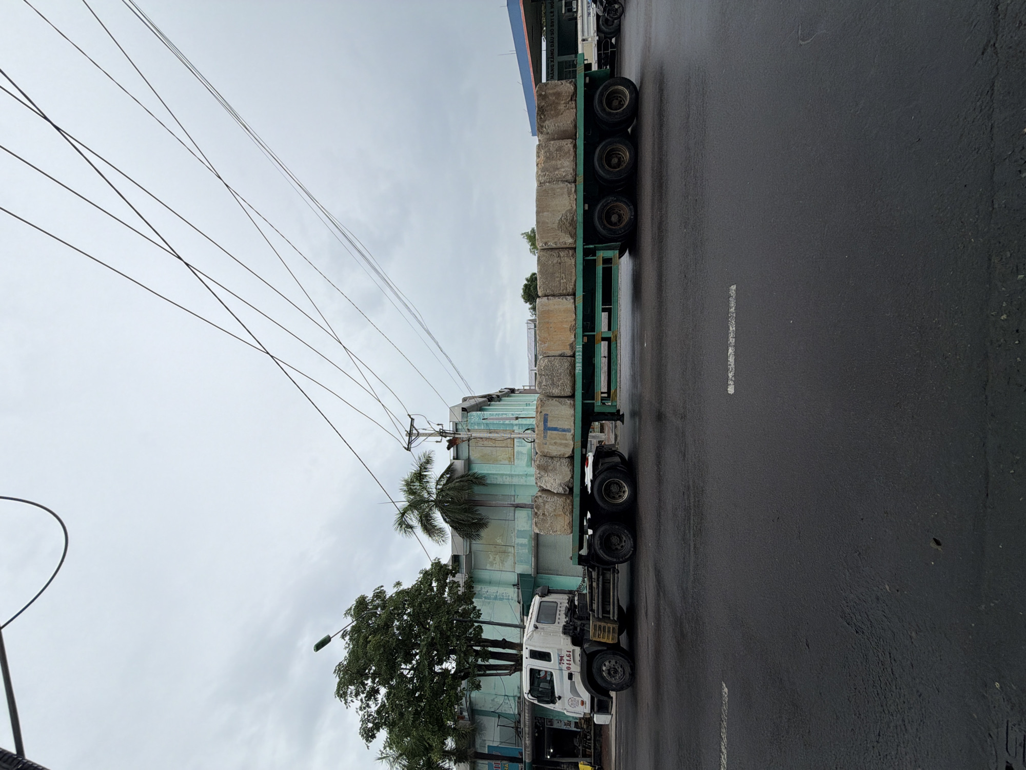 A white semi-trailer truck is transporting large, rectangular concrete blocks on a flatbed trailer. The truck is on a wet, asphalt road, possibly in an industrial or commercial area of Nha Trang, Vietnam, given the context of the city. The sky is overcast, suggesting a cloudy or rainy day. In the background, there are buildings, trees, and overhead power lines, which are common features in urban Vietnamese landscapes. The blocks appear to be roughly uniform in size and are stacked closely together on the trailer. There are no people visible in the immediate vicinity of the truck or the cargo. The scene depicts a moment of transport of construction materials.