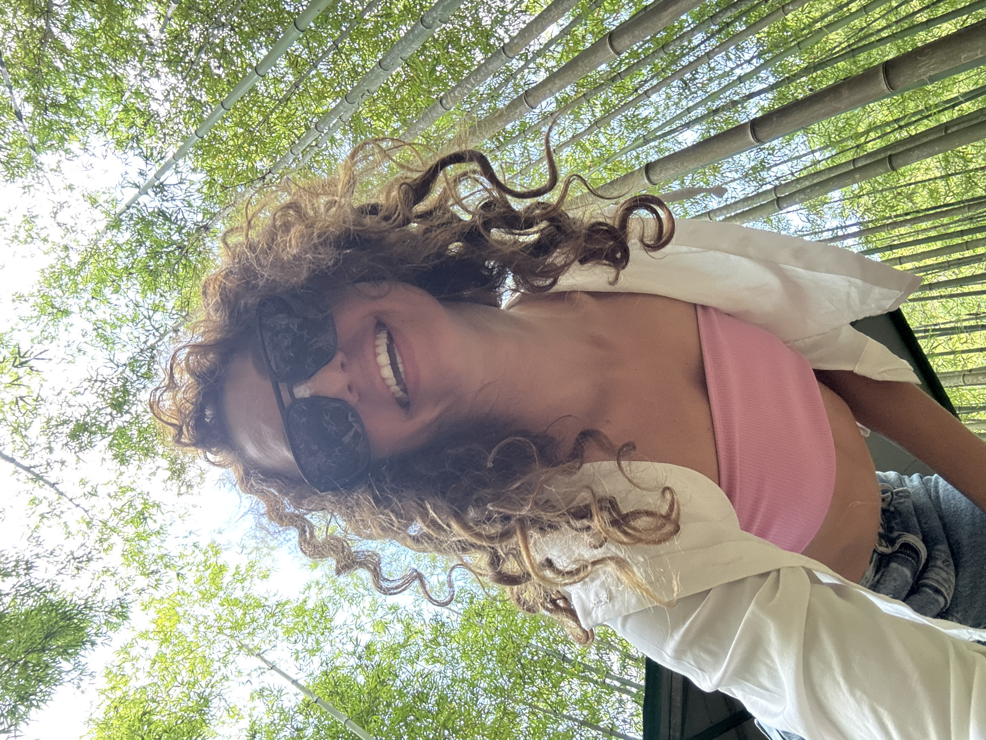 A woman with curly brown hair and sunglasses is seen from a low angle, likely holding a phone and taking a selfie or video. She is wearing a pink top under a white shirt, and some blue denim is visible on her lower right. She is smiling. The background appears to be a bamboo forest with light filtering through the dense canopy. This is consistent with the area of Púng Luông, Vietnam. Bamboo stalks extend vertically overhead, and foliage is visible through the gaps. The sunlight is bright, suggesting it is daytime.