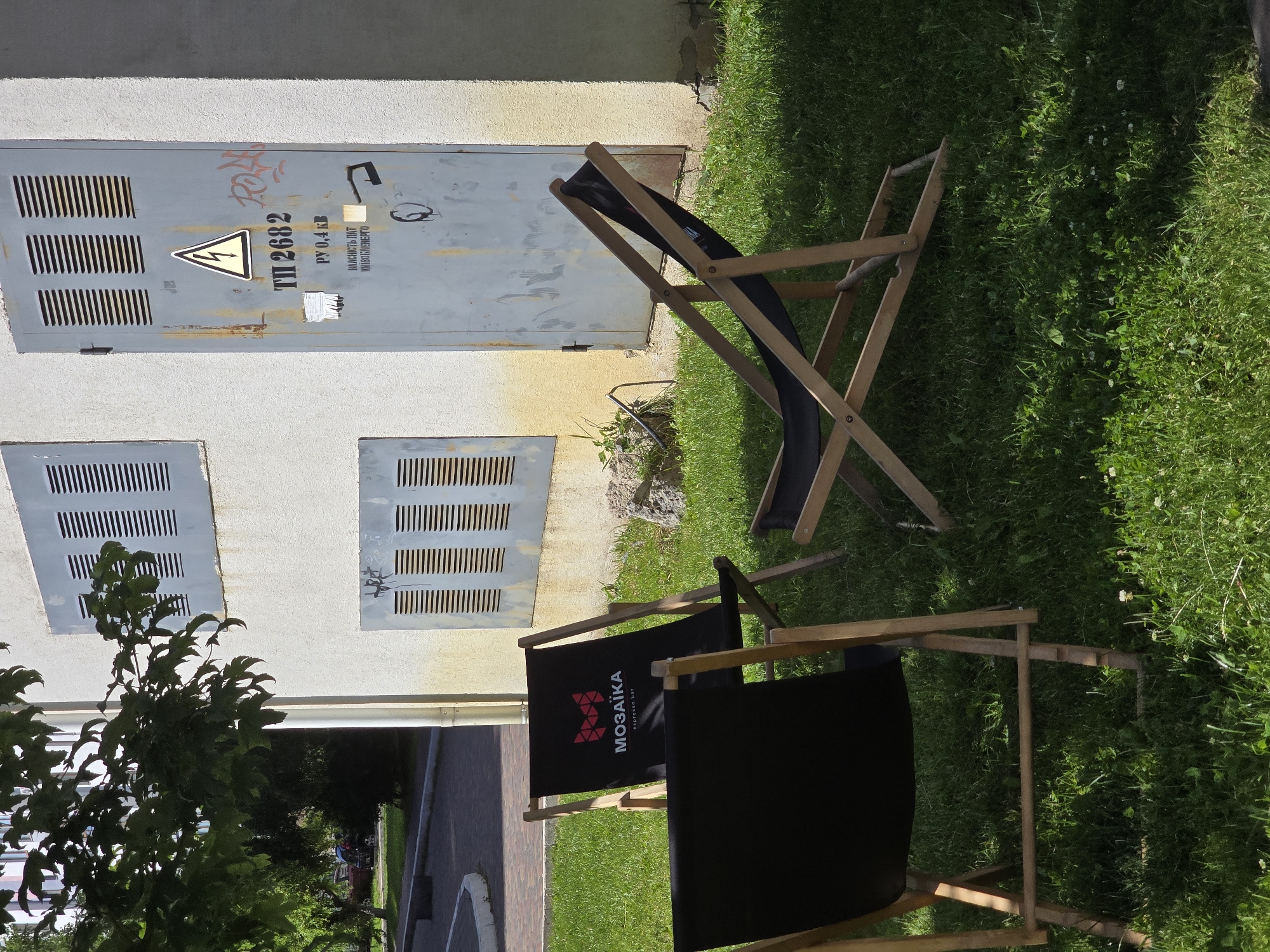 This is a medium shot, eye-level, outdoor photograph taken on a sunny day. In the foreground, on a patch of green grass with some small white flowers, are three wooden and black fabric deck chairs. One chair in the center has a red and white logo with the word "МОЗАЙКА" (MOSAIKA) and "espresso bar" printed on it. The chair in the foreground on the left is solid black. A third chair is partially visible on the right. Behind the chairs is a weathered, light gray building with several metal grates and a metal door. The door has a yellow triangle with a lightning bolt symbol and Ukrainian text indicating it's an electrical hazard, along with other markings. The building is stained with yellow and has some graffiti. To the left of the building, part of a leafy green tree branches into the frame, casting shadows. In the background, a paved area and some trees are visible, suggesting an urban or suburban setting. This scene is located in Petropavlivska Borshchahivka, Ukraine.