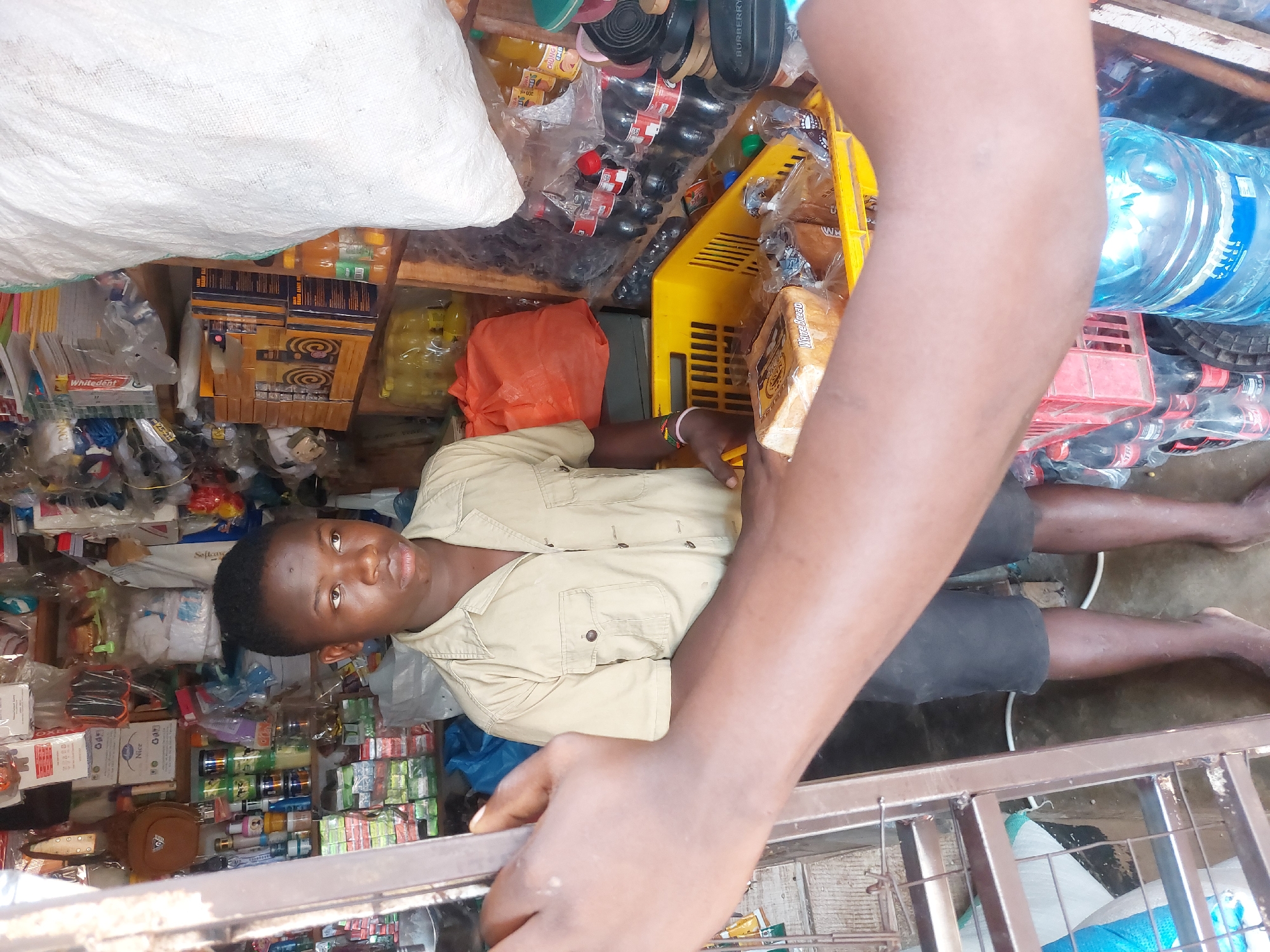 A man wearing a khaki shirt and black shorts lies in what appears to be a small shop, potentially in Dar es Salaam, Tanzania. The shop is filled with various goods, including drinks, food items, and toiletries. The man appears to be resting or lying down, with his legs extending along the floor. The environment is crowded with merchandise. A person is holding onto a metal frame of what appears to be a window to the establishment.