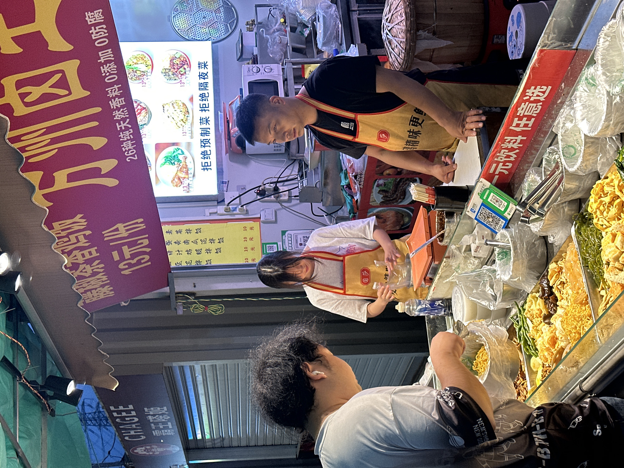 Here is a detailed description of the image:
**Scene Overview:**
The image appears to be taken at a street food stall in Jiangjin, China, showcasing the preparation and sale of various food items. The shot is composed from the customer's point of view.
**People:**
1. **Customer:** A person with curly black hair wearing a white t-shirt is seen holding a plastic bag and using tongs to select food from the display.
2. **Food Stall Workers:** There are two food stall workers: one male and one female. Both are wearing aprons over their clothes. The man is standing behind the counter, interacting with the food items, and the woman is also working behind the food display area.
**Objects:**
1. **Food Display:** A wide display case filled with various types of food, including noodles, vegetables, and what appear to be fried snacks or ingredients.
2. **Signage:** Various signs are present, with Chinese text indicating prices (e.g., "13元/份," "10元"), promotional messages ("26种纯天然香料 0添加 0防腐"), and business names ("CHAGEE").
3. **Food Preparation Area:** The food stall includes a microwave, containers, and other equipment typical of a food preparation area.
4. **Payment System:** A QR code for mobile payment is visible, likely for payment transactions.
5. **Beverages:** There's signage indicating beverages available ("一元饮料 任意选").
**Actions & Interactions:**
1. **Customer Interaction:** The customer is actively selecting food items using tongs.
2. **Worker Activity:** The workers are preparing and serving food, attending to the customer's order.
**Environment:**
1. **Food Stall Setup:** The environment is a typical outdoor food stall setup with signs, display cases, and food preparation equipment.
2. **Lighting:** The scene appears to be well-lit, possibly with artificial lighting due to the time of day.
3. **Text and Messaging:** The Chinese text on the signs provides further details about the food ingredients and marketing messages.
**Summary:**
This photograph shows a bustling food stall environment in Jiangjin, China, with customers selecting food and stall workers preparing and serving. The scene captures the essence of local street food culture and commercial activity.