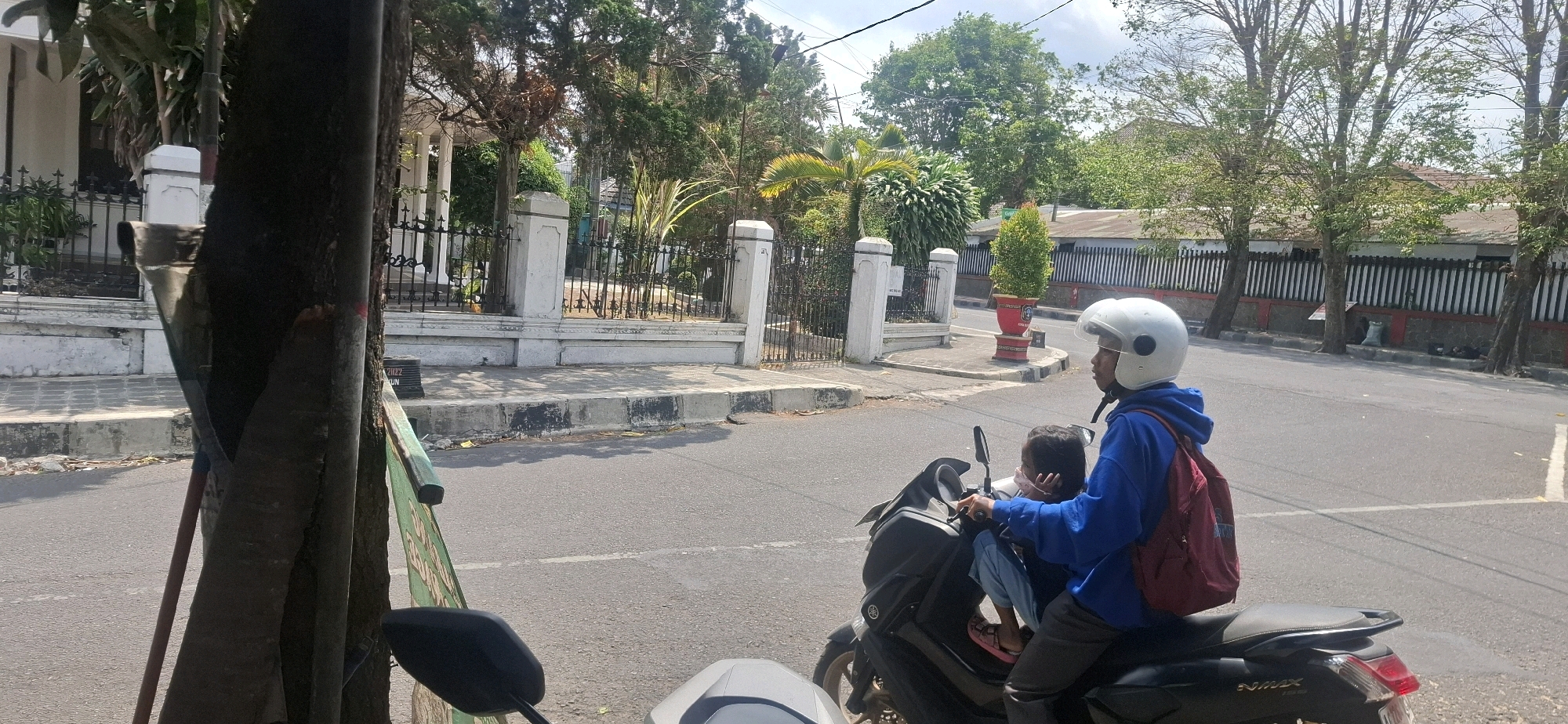 A man in a blue hoodie and white helmet rides a dark-colored scooter with a young girl in the passenger seat, who is wearing a pink mask and blue pants. They are traveling on a paved road, passing a sidewalk and a white fence with black vertical bars. Behind the fence are lush green trees and a building with a white facade and columns. The scene is set on a bright, sunny day in Blitar, Indonesia. A large tree trunk occupies the left foreground of the image, partially obscuring the view of the road and sidewalk.