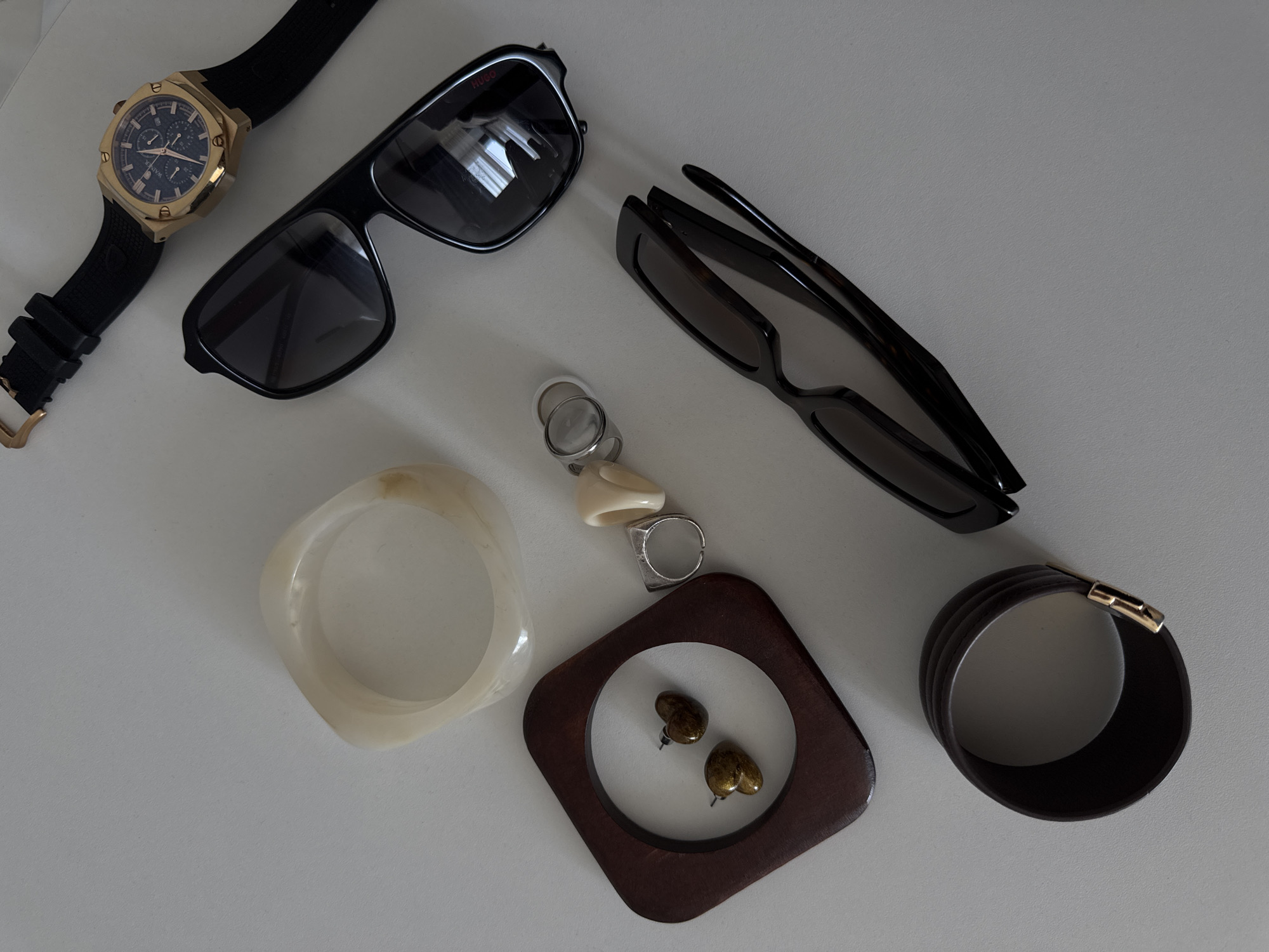 This image presents a flat lay arrangement of various personal accessories on a plain, light grey surface, indicative of an indoor setting within the city of Sochi, Russia.
From top to bottom and right to left, the items are:
1. A men's gold-toned chronograph watch with a dark blue dial and a black rubber strap, secured by a gold-toned buckle. The brand "WARBURTON" is partially visible on the dial.
2. A pair of black, rectangular-framed sunglasses with dark tinted lenses.
3. An off-white, irregularly shaped square or oval bangle, appearing to be made of a smooth, translucent material.
4. A collection of rings and earrings:
* Two silver-toned rings, one a plain band, the other an adjustable band.
* A ring featuring a cream-colored, pointed, possibly bone or horn-like element.
* Two small, heart-shaped stud earrings with a bronze-gold metallic finish, positioned inside a dark brown wooden square frame with a circular cutout.
5. A second pair of dark-framed sunglasses, folded, with a slightly more angular or tapered design than the first pair.
6. A dark brown, possibly leather, cuff bangle with a ribbed or layered design, featuring a small gold-toned clasp.
The objects are scattered across the surface without a distinct pattern, illuminated by an overhead light source that casts subtle shadows. No individuals are present in the scene, and no actions are occurring.