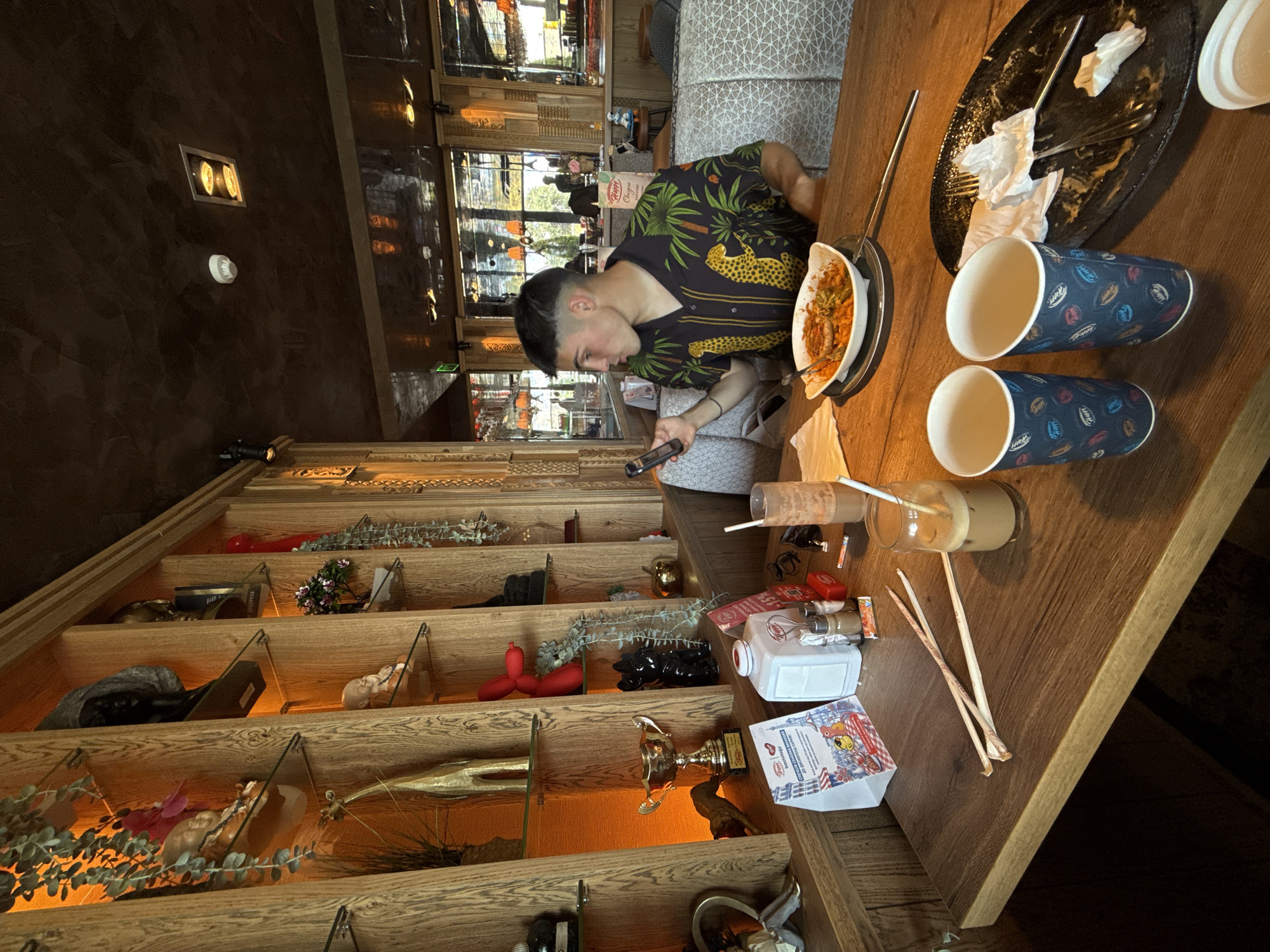 The image captures an indoor scene, likely a restaurant or culinary center in Radinovo, Bulgaria, oriented vertically (rotated 90 degrees counter-clockwise). On the right, a young man with a short, faded haircut is seated at a wooden table, engrossed in his smartphone. He wears a dark short-sleeved shirt patterned with green palm trees and yellow leopards.
On the table in front of him are the remnants of a meal in an oblong white dish, a dirty black plate with used napkins, and two blue paper cups with a colorful leaf pattern. An empty glass jar with a straw suggests a finished beverage, and various small items like sachets and a white bottle (possibly labeled "Happy Original") are also present. The background behind the man shows large windows with reflections of an outdoor scene, suggesting it is daytime.
To the left, a multi-tiered wooden shelf unit extends vertically, filled with a diverse collection of decorative objects. These include a golden trophy, small statues (such as a baby doll figure), artificial plants, bottles, a red balloon animal, and a black panther figurine. The shelves are warmly lit from below. A prominent white sign with a red border on one shelf clearly reads "РАДИНОВО" (Radinovo), "КУЛИНАРЕН ЦЕНТЪР" (Culinary Center), and "BULGARIA", confirming the location as a culinary center in Radinovo. Another small card visible on a shelf has "HAPPY CLUB" written on it. The walls are a dark, textured material, and ceiling lights are visible. The overall ambiance appears casual and well-decorated.
