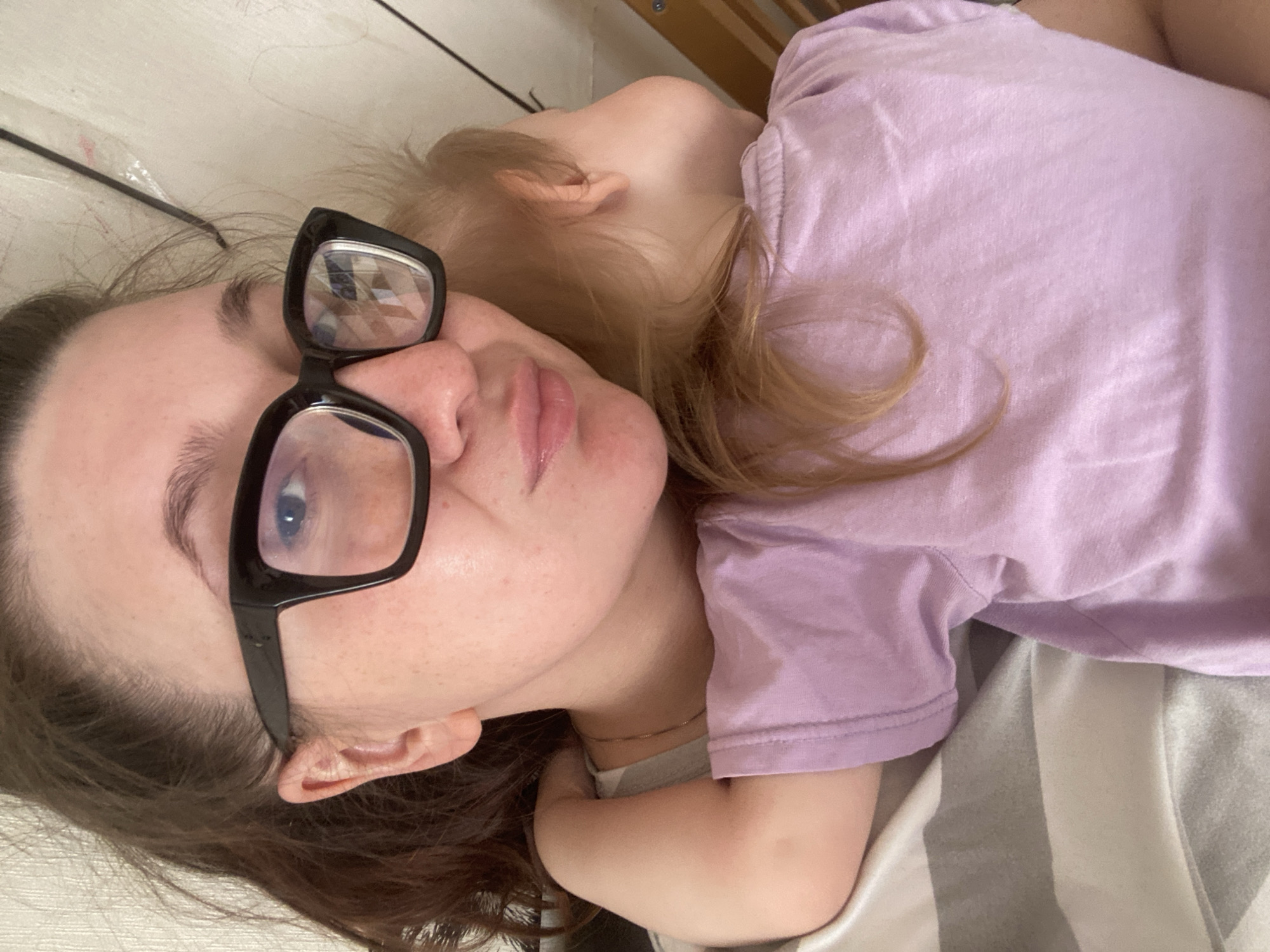 A woman wearing black-framed eyeglasses is lying on a striped surface next to a young child. The child is wearing a purple shirt. The woman's location is specified as Chaikovsky, Russia.