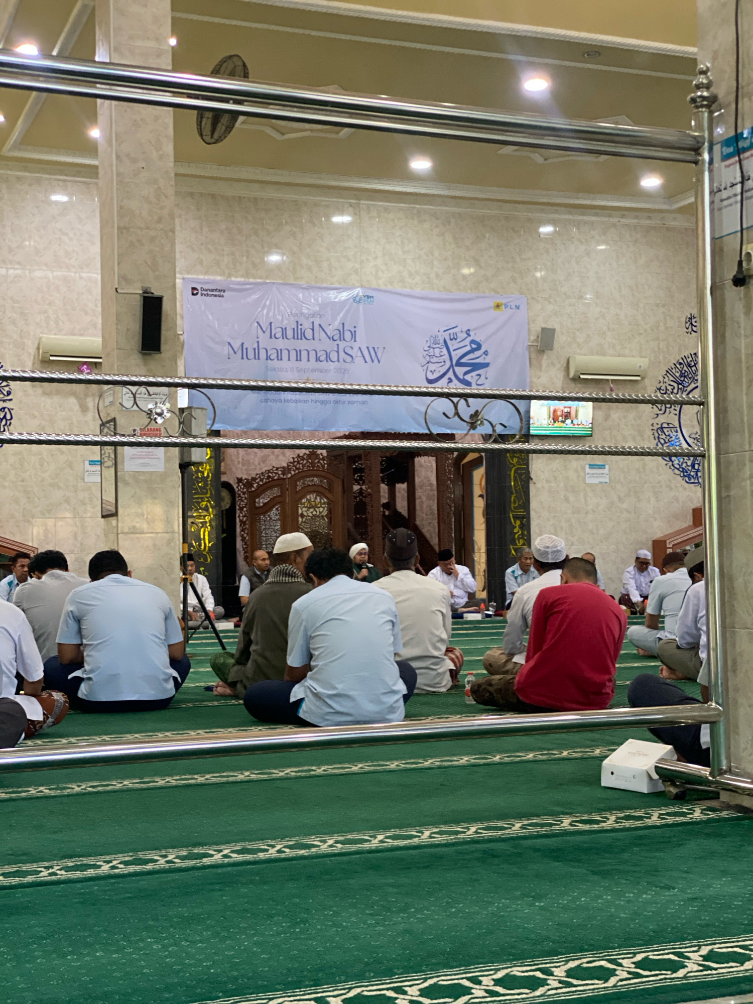 The image captures an indoor gathering, likely a religious event, taking place within a mosque. In the foreground, several men are seated on a green carpet, facing away from the camera. They are dressed in casual and traditional attire, with some wearing prayer caps. The carpet has decorative white patterns. A metallic railing runs horizontally across the middle of the frame, partially obscuring the view of the congregants and the area beyond.
In the background, a large banner is displayed on the wall, bearing Indonesian text that translates to "Commemoration of the Birthday of the Prophet Muhammad SAW". Below this, a smaller line of text indicates "Tuesday, 8 September 2026". Arabic calligraphy, likely verses from the Quran or religious salutations, is visible on the wall. A ceiling fan is mounted on the ceiling, and lighting fixtures illuminate the space. An air conditioning unit is also present on the wall. Further back, additional people are visible, some seated, and others appearing to be standing or moving. A television screen displays an image of people, possibly an ongoing broadcast related to the event. The architecture suggests a formal building, with marble-patterned walls and ornate wooden doorways visible in the deeper background. The overall scene depicts a solemn and communal religious observance. The event is identified as taking place in Banjarbaru, Indonesia, indicating the specific location of this mosque and gathering.