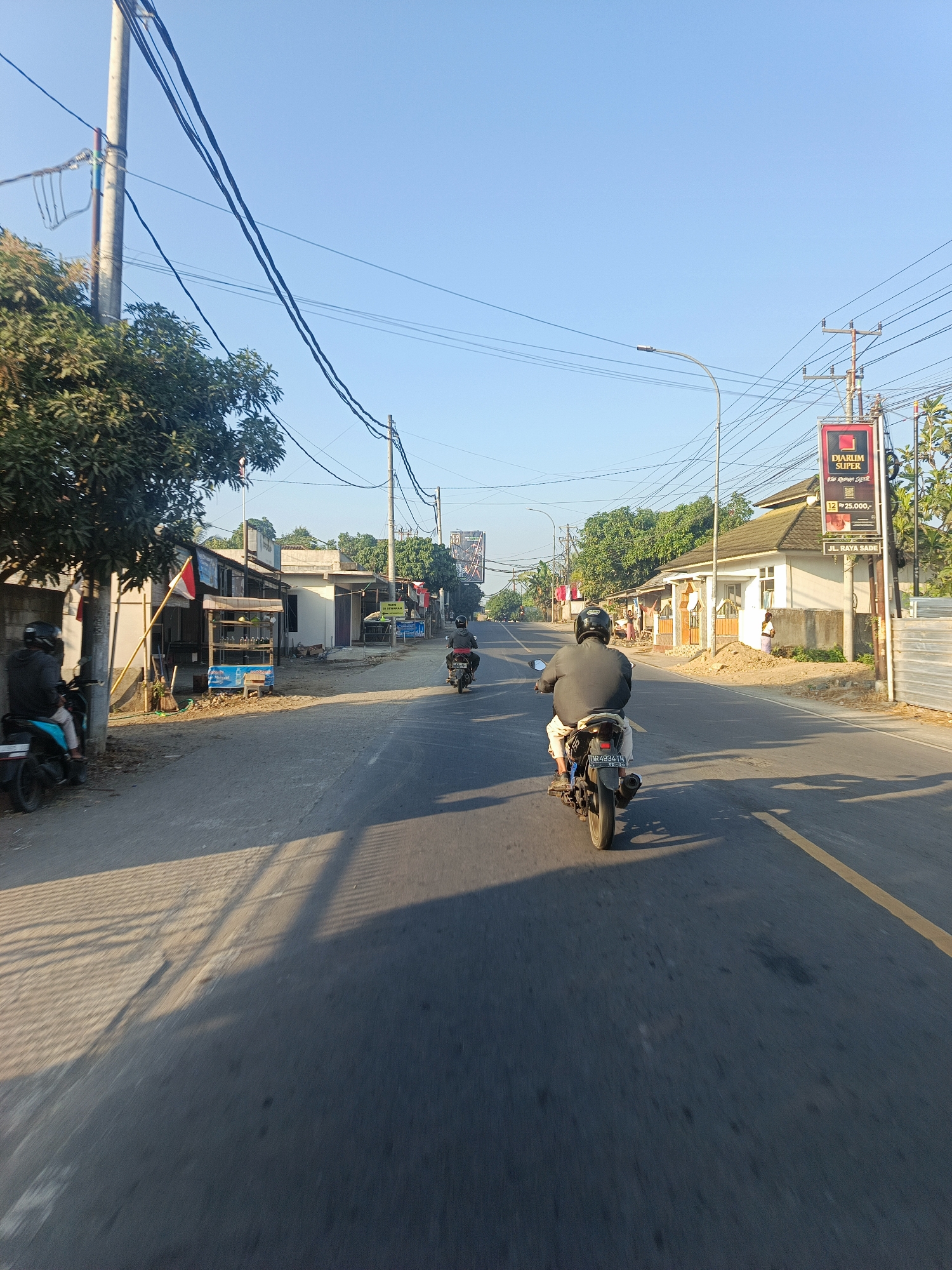 The image depicts a street scene in Sengkol, Indonesia, under a clear blue sky. The perspective is from the point of view of someone riding a motorcycle, looking forward. A man wearing a dark jacket and helmet is riding a motorcycle in the foreground, his back to the camera. The motorcycle has a license plate visible.
To the left of the road, there are trees and a small building with an Indonesian flag displayed. A person on a blue and black motorcycle is parked on the left side of the road. Further down the road, other motorcycles are visible, as well as more buildings and trees lining the street. A prominent billboard for "Djarum Super" is on the right side of the road, along with power lines and utility poles. Some people are visible near the buildings on the right side of the street. The road is paved with asphalt and has a yellow line marking. Sunlight casts long shadows across the road, indicating it is likely early morning or late afternoon.