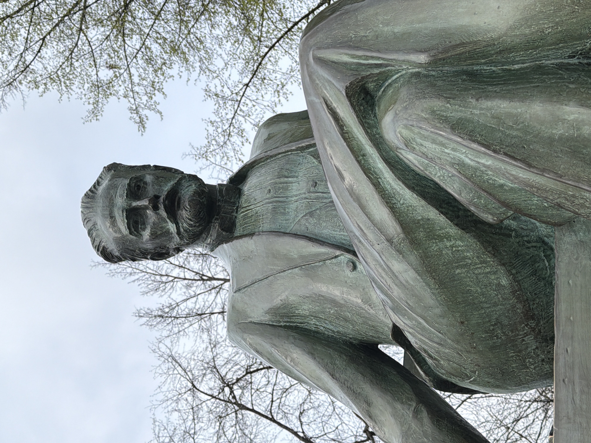 The image shows a bronze statue of Pyotr Ilyich Tchaikovsky, a famous Russian composer, in Votkinsk, Russia, his birthplace. The statue depicts Tchaikovsky in a formal suit with a long coat draped over his shoulder. Bare tree branches are visible in the background against a cloudy sky. The camera is positioned low, looking up at the statue.