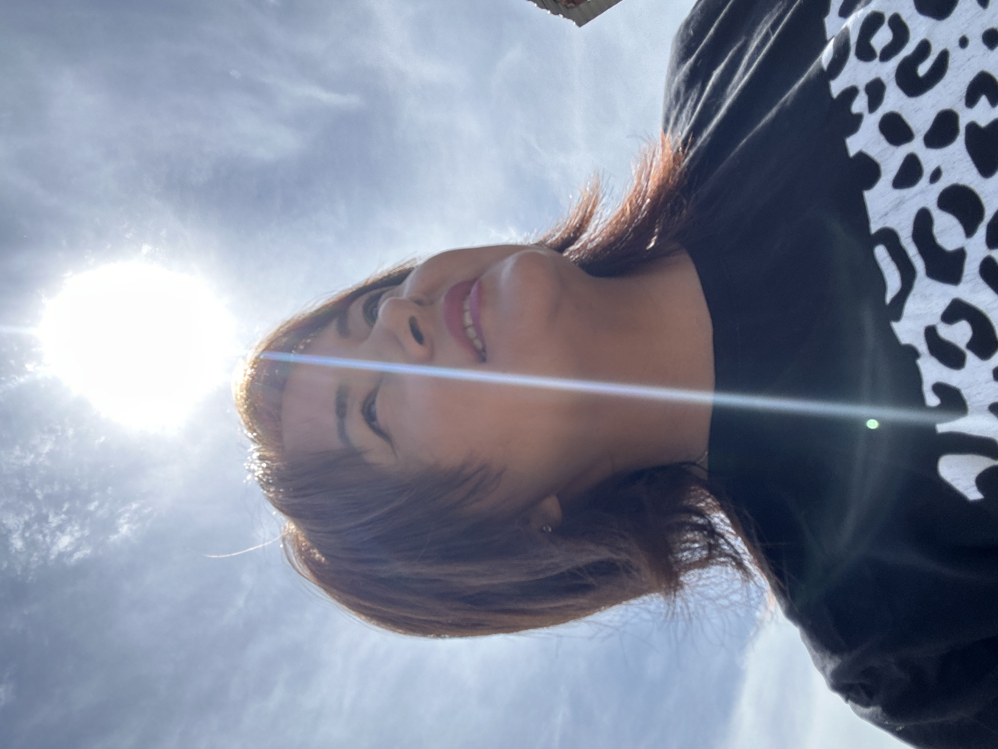 A woman is captured in a low-angle shot, looking upwards towards a bright, hazy sun. The sun is the dominant light source, creating lens flare and washing out parts of the sky, which is filled with scattered clouds. The woman has shoulder-length, reddish-brown hair with layers that frame her face. She is wearing a black t-shirt with a white leopard print pattern on the lower portion. Her facial features are visible, including her eyes, nose, and lips, which appear to be painted with pink lipstick. The background is the sky, suggesting an outdoor setting. There are no other discernible people or objects in the immediate vicinity. The image appears to be a selfie or a photograph taken from a low perspective.
