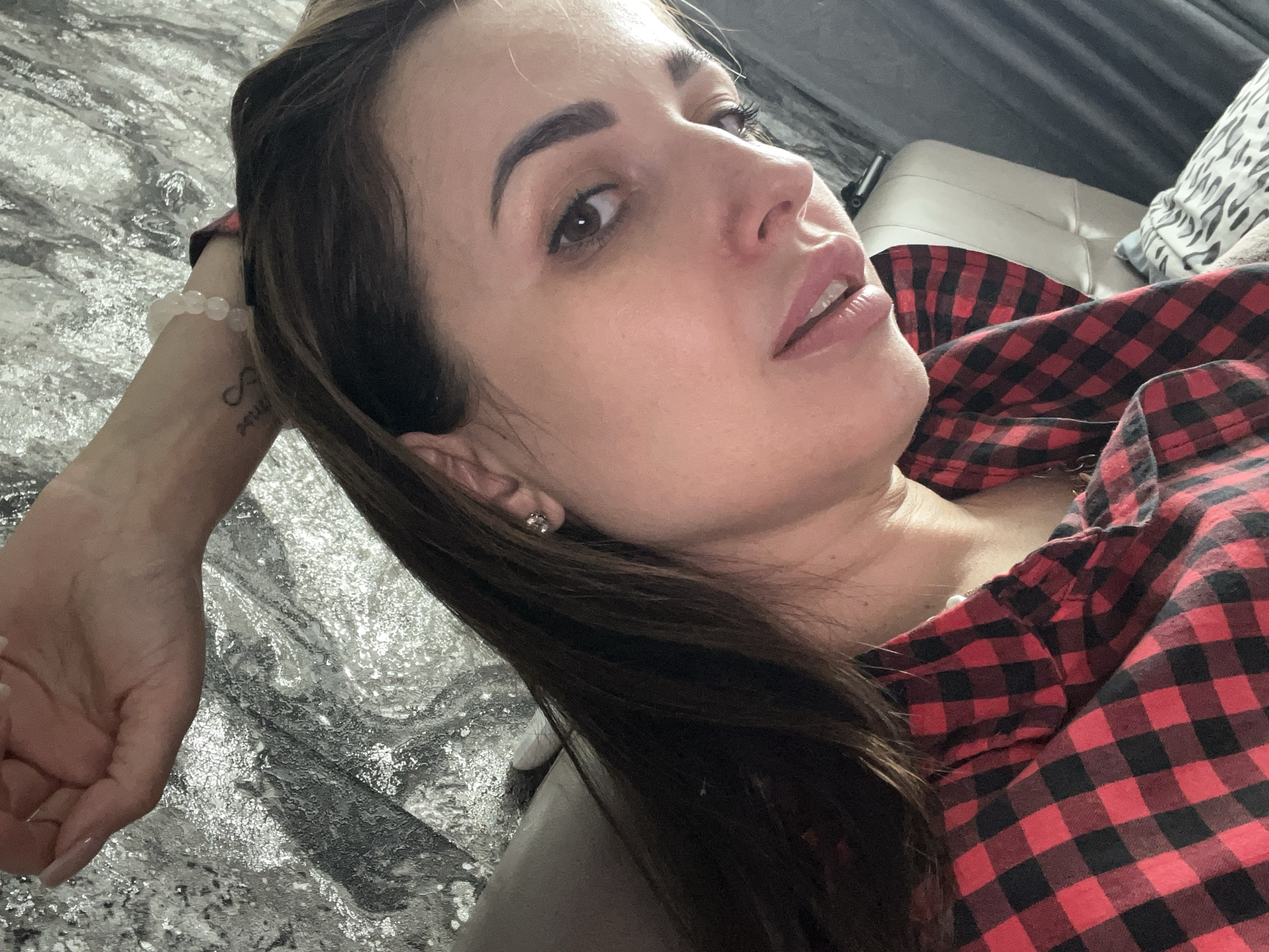 A woman lies at an angle, her head closer to the viewer. She has brown hair, fair skin, and brown eyes, and wears a red and black checkered shirt. A silver patterned surface is visible to her left, and her left arm rests on it, adorned with a beaded bracelet and a tattoo. In the upper right background, there are glimpses of a white sofa and a patterned cushion.