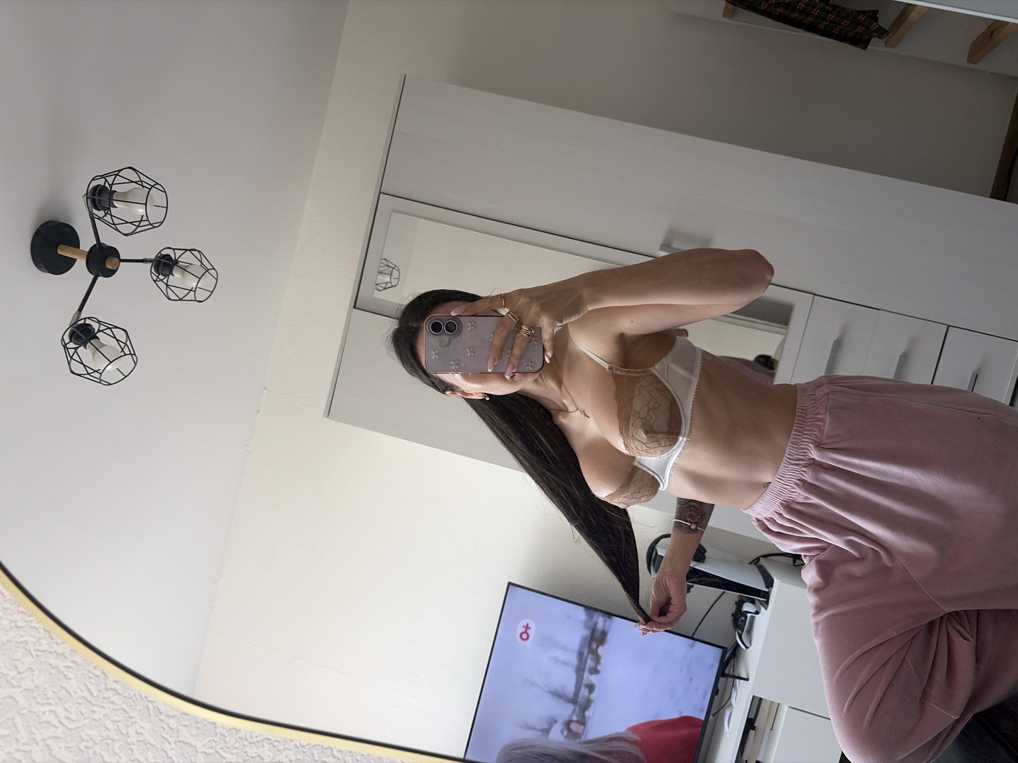 Here is a detailed description of the image:
**Scene:** A reflection captured in a mirror shows a young woman taking a selfie in what appears to be a bedroom.
**People:** The primary subject is a woman, who is holding up a smartphone to take a picture of herself in the mirror.
**Objects:**
* **Mirror:** A large mirror with a gold frame.
* **Smartphone:** A smartphone, presumably used for taking the selfie. The back of the phone has a floral design.
* **Clothing:** The woman is wearing a bra and pink sweatpants.
* **Television:** In the background, there is a television showing a snowy scene.
* **Furniture:** Part of a white wardrobe with drawers is visible.
**Actions:** The woman is taking a selfie in the mirror. She has long hair which she is holding at the end.
**Interactions:** The woman is interacting with her phone to take a picture.
**Environment:** The setting seems to be the woman's bedroom. The background includes a television, furniture and the bedroom walls.