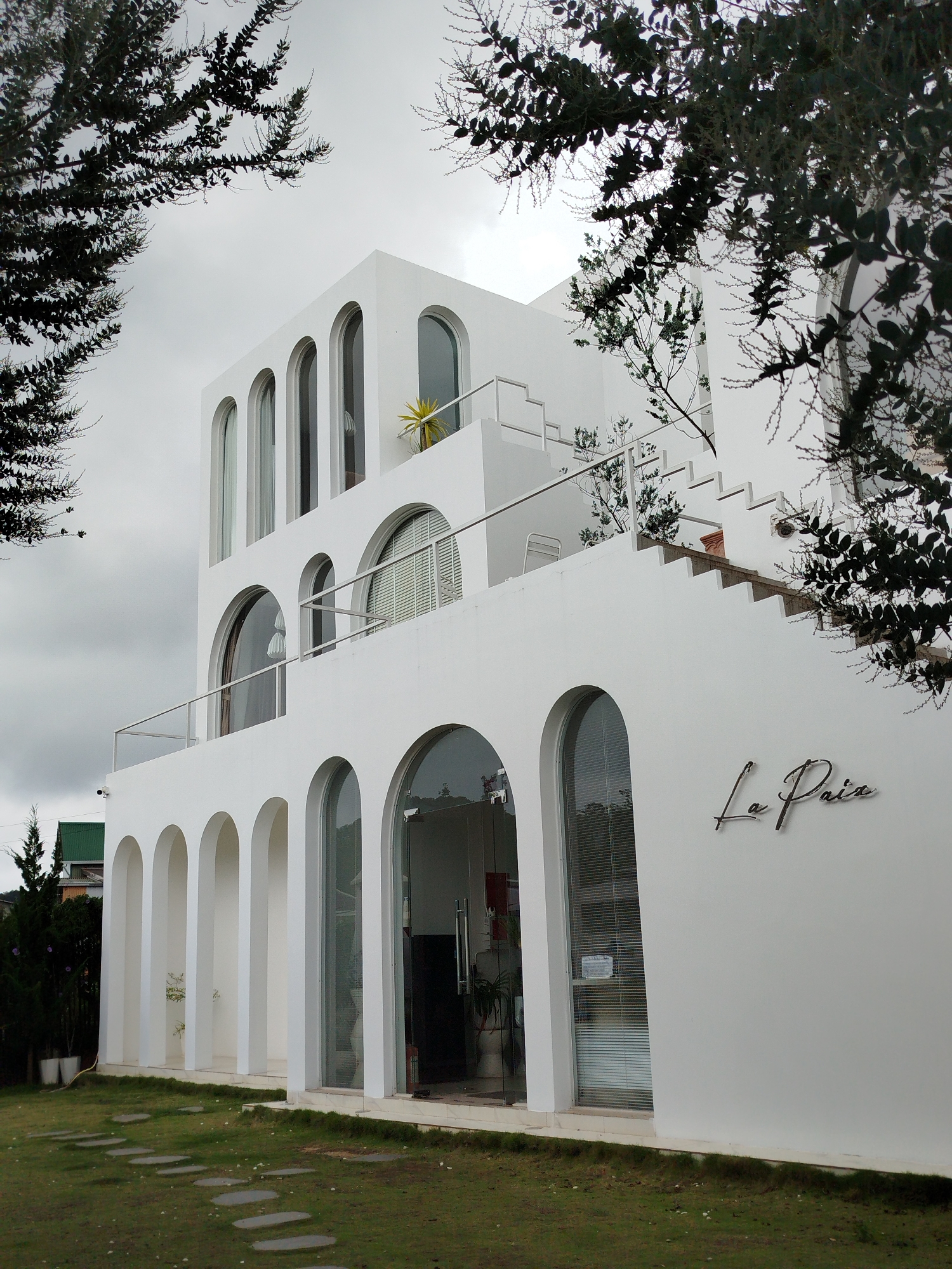 A white, multi-story building with arched windows and doorways, in the city of Da Lat, Vietnam, is captured from a low angle. The building has a modern architectural style, featuring clean lines and a minimalist aesthetic. The facade is adorned with the script "La Paix" in what appears to be brushed metal.
The building is partially obscured by the dark foliage of trees in the foreground, framing the upper portion of the image. The sky is overcast, contributing to a diffused light that casts soft shadows.
In front of the building, a pathway of flat, grey stepping stones leads across a patch of green grass. The grass appears well-maintained, with some scattered white wildflowers visible. To the left of the building, a small evergreen tree stands, and further in the distance, a glimpse of a green-roofed structure can be seen.
The lower level of the building features a series of arched openings, some of which are filled with large glass panes that reflect the surroundings and reveal glimpses of the interior. A clear glass door is visible on the right side of the ground floor. The upper floors have more arched windows, and a balcony with a glass railing is present on the second level. A white staircase ascends the right side of the building.
No people are visible in the image. The scene depicts an exterior view of the building and its immediate surroundings, suggesting a serene and tranquil setting.