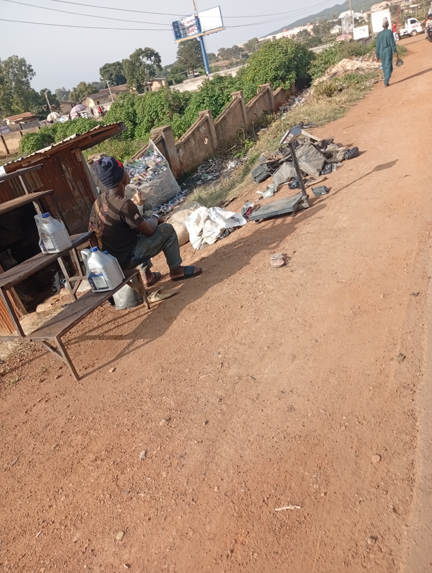 This is a full, eye-level, outdoor, medium shot of a street scene in Jos, Nigeria, where a person is sitting on a metal bench. The person is wearing a dark beanie and a dark t-shirt with an emblem on the back, dark pants, and flip-flops. They are sitting on the bench, facing left, with their hands on their lap. Next to the person on the bench are two large white plastic containers. The bench is part of a small, makeshift stall made of corrugated metal sheeting. Behind the stall, a large, woven bag filled with plastic bottles is visible. The ground is a reddish-brown dirt. To the right of the stall, on the dirt road, is a pile of what appears to be discarded electronics and metal parts, including a large weighing scale. Further down the road, a man in a green robe and hat is walking away from the camera. In the background, there are buildings, trees, and a billboard. The sky is clear and bright.