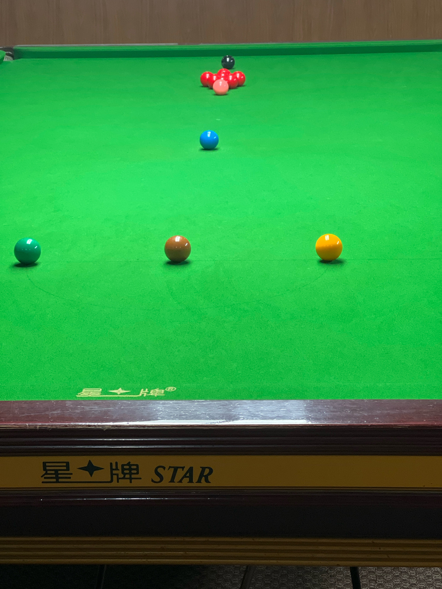 The image depicts a snooker table with several balls arranged on its green felt surface. In the foreground, there are three balls: a green one on the left, a brown one in the center, and a yellow one on the right. Further up the table, there is a single blue ball. Near the top of the table, a cluster of red balls and one pink ball are arranged, with a single black ball behind them. The table has a wooden edge and a yellow stripe with Chinese characters and the word "STAR" written on it. The background appears to be a wooden wall. The scene is indoors. There is no indication of the city of Attock, Pakistan.