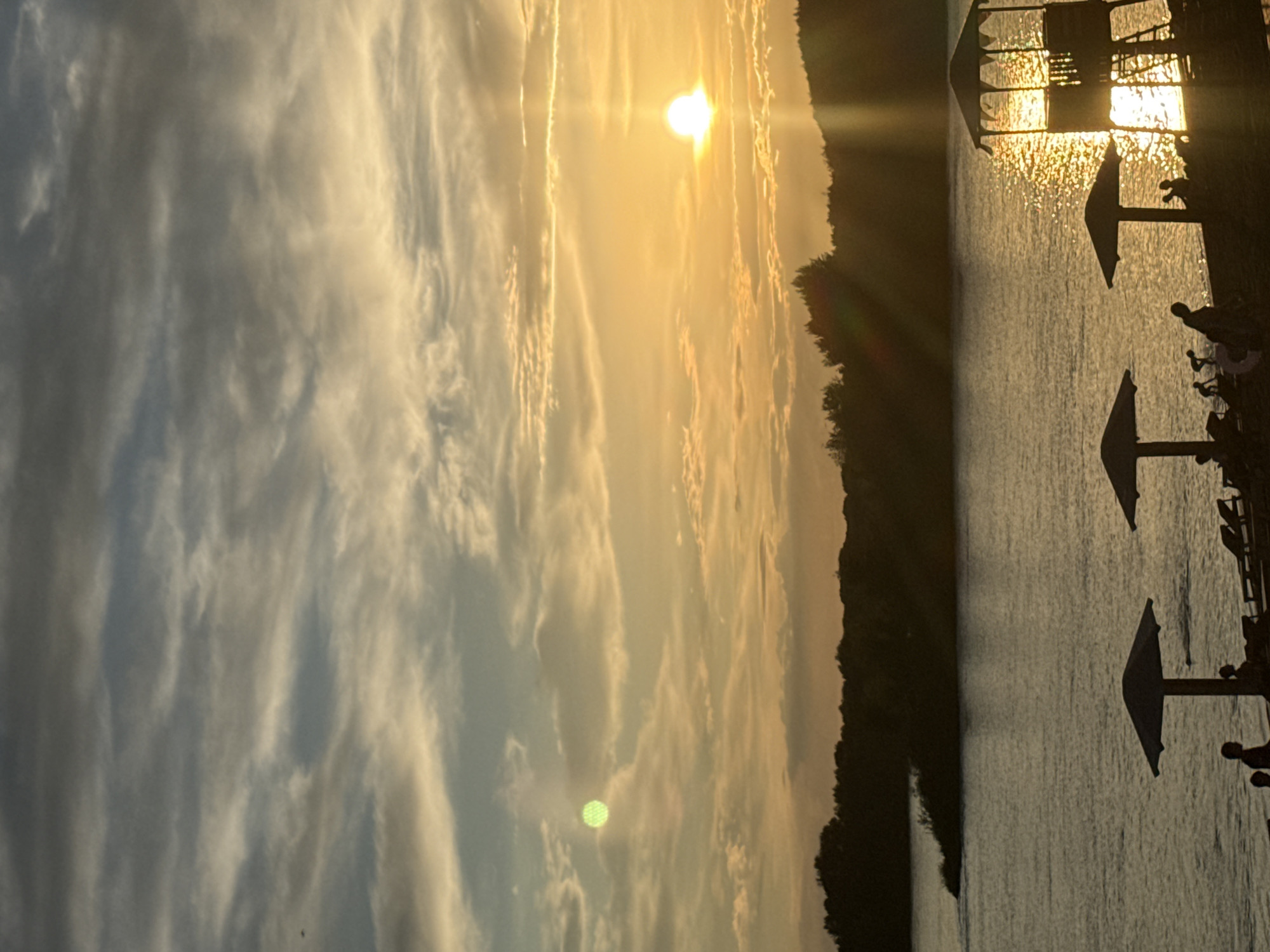 This image depicts a sunset over a body of water, likely the Irtysh River, in Pavlodar, Kazakhstan. The sky is filled with a dramatic display of clouds, with the setting sun casting a warm, golden glow. The sun, a bright orb, is positioned in the upper right quadrant of the image, its rays reflecting on the water's surface.
In the foreground, a beach area is visible, silhouetted against the vibrant sky. Several beach umbrellas are set up, providing shade. The silhouettes of people can be seen near the water's edge and under the umbrellas, suggesting a leisurely gathering. To the right, a lifeguard tower stands prominently, its structure also in silhouette. The dark, treed shoreline of a distant island or riverbank forms the middle ground, separating the water from the sky. The overall scene conveys a sense of tranquility and the end of a summer day.