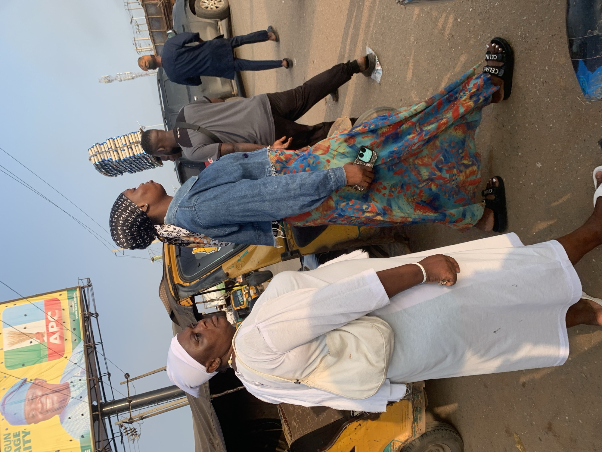 In Shagamu, Nigeria, a group of people are seen near a yellow auto-rickshaw. A woman in a white dress and headscarf stands on the left, carrying a cream-colored bag. To her right, a woman in a denim jacket and a vibrant floral skirt stands looking away, holding a phone. Behind them, another woman with a patterned headscarf is partially visible. Several men are in the background, one wearing a grey t-shirt and another in a dark blue outfit. A large billboard featuring a politician and the APC logo is visible in the upper left corner. The environment appears to be a street with vehicles and a telecommunications tower in the distance. The overall scene depicts a busy urban setting.