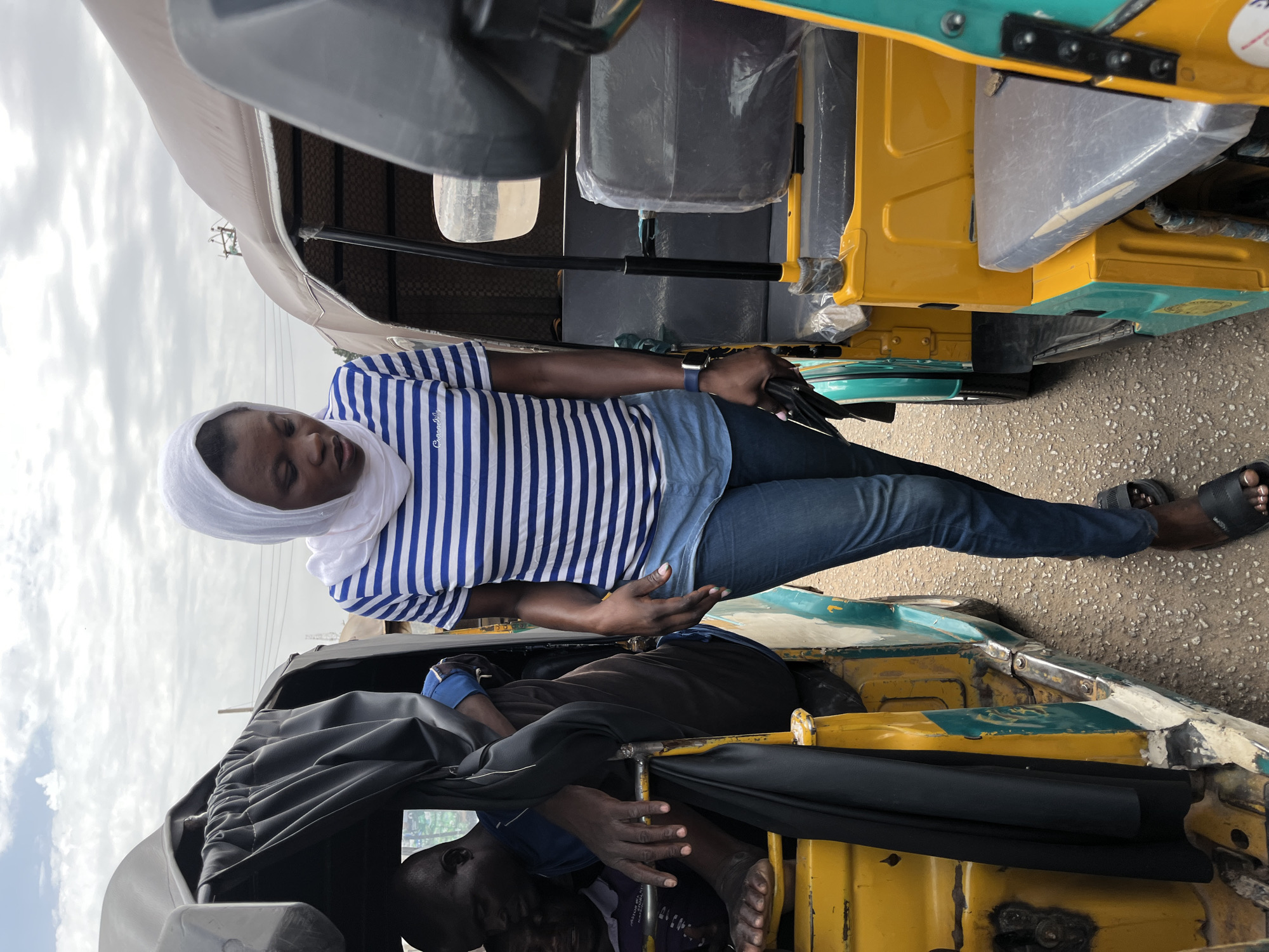 A woman in a white headscarf and a blue and white striped shirt sits in a yellow and green auto rickshaw (tuk-tuk) on a paved street in Gura Top, Nigeria. Another individual is partially visible in the lower part of the vehicle, lying down, covered with a black material. A second auto rickshaw is parked closely beside the first. The sky is overcast.