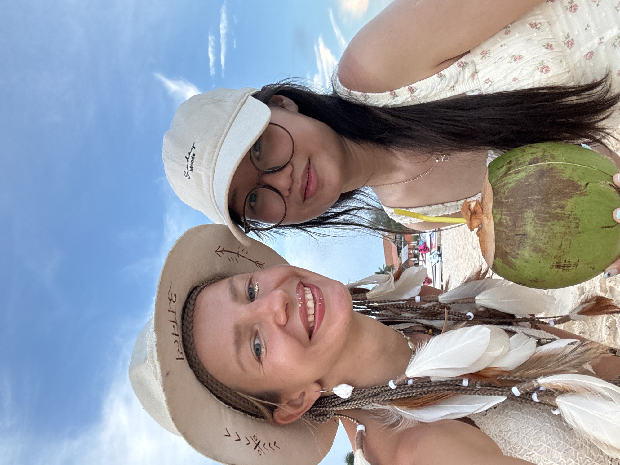 Two young women are taking a selfie on a beach in Mũi Né, Vietnam. The woman on the left is wearing a wide-brimmed, light-colored hat with brown designs. Her hair is styled in braids adorned with white feathers and beads. She is smiling and has glitter on her face. The woman on the right is wearing a light-colored baseball cap with "Sunday Moods" written on it, and glasses. She has long, dark hair. She is holding a whole coconut with a straw. Both women are wearing light-colored dresses. The background shows a sandy beach with some buildings and lounge chairs visible further in the distance, under a bright blue sky with some clouds. The overall scene suggests a relaxed, tropical vacation.