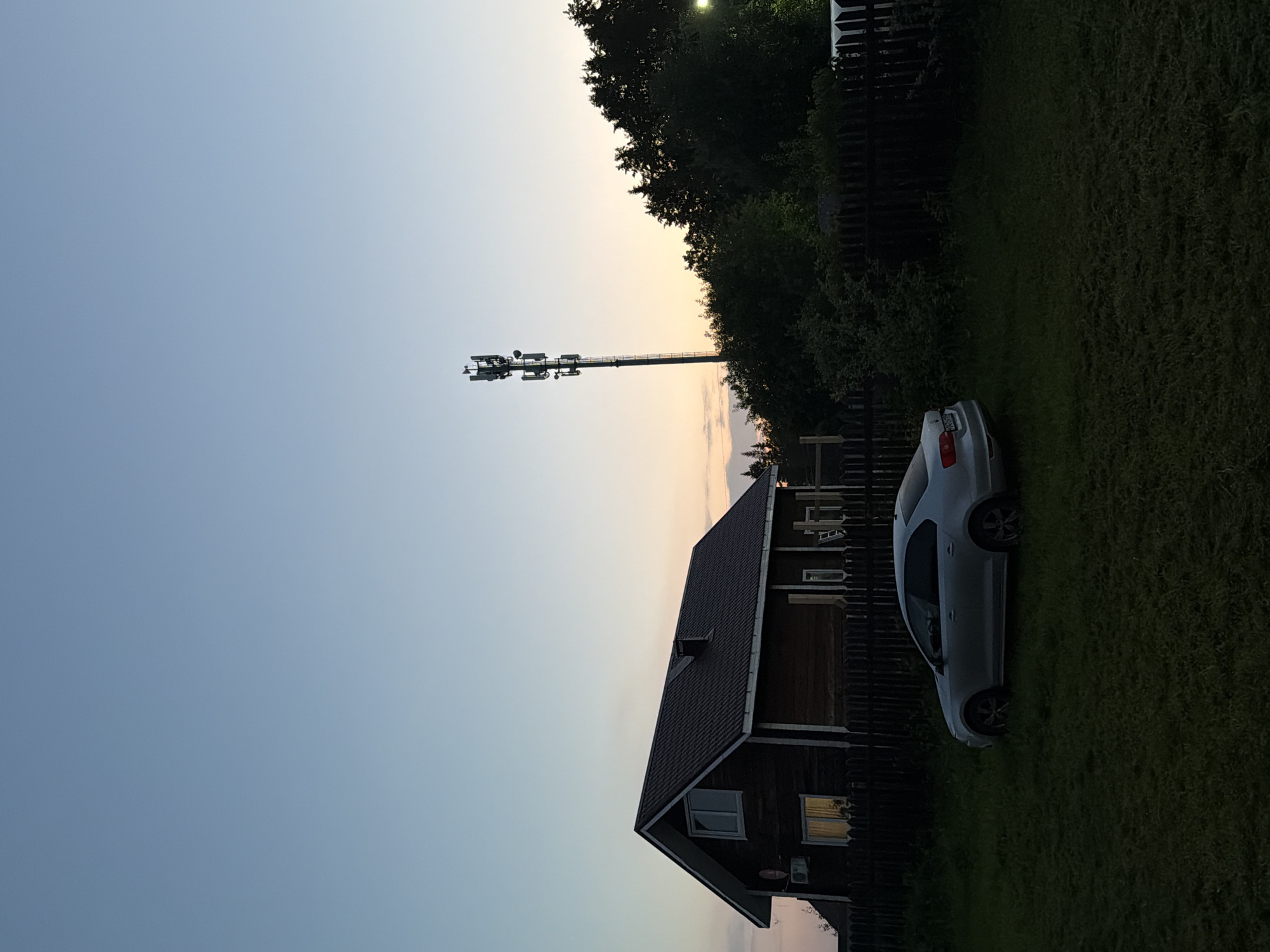 The image depicts an outdoor scene at dusk, likely in Kultaevo, Russia, given the context. In the foreground, a silver sedan is parked on a grassy lawn. To the left of the car is a dark wooden house with a pitched roof and windows. A dark wooden fence runs along the front of the property. In the background, a tall cell tower with multiple antennas stands against the pale orange and blue twilight sky. A dense line of dark green trees forms a backdrop to the cell tower and extends across the middle ground. The overall lighting suggests it is either early evening or late evening.