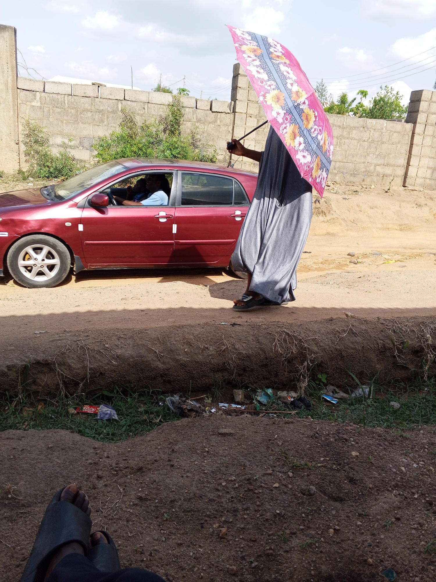 This daytime photograph, taken in Adonkolo, Nigeria, depicts an outdoor scene along an unpaved, dusty road. A red sedan car, possibly a Toyota Corolla given its design, is visible on the road. A person wearing a light blue shirt is seated in the driver's seat, their upper body and arm visible through the car window.
Standing beside the car on the right, another individual is largely obscured by a flowing grey garment and a vibrant pink umbrella. The umbrella, adorned with prominent yellow, white, and blue floral patterns, is held aloft, likely providing shade from the sun. The person's feet, clad in black sandals, are visible at the bottom of their garment.
In the background, a low concrete block wall spans the scene, with green shrubs and sparse vegetation growing along its base and behind it. The sky above is bright with scattered clouds, indicating sunny weather.
The foreground of the image features a dirt embankment or ditch, where patches of green grass grow amidst scattered litter, including plastic bags and a red and white wrapper with the word "Trixie" partially visible. The photograph is taken from a low vantage point, with the photographer's feet, also wearing black sandals, appearing in the very bottom left corner of the frame. The scene portrays a typical street environment in a warm climate.