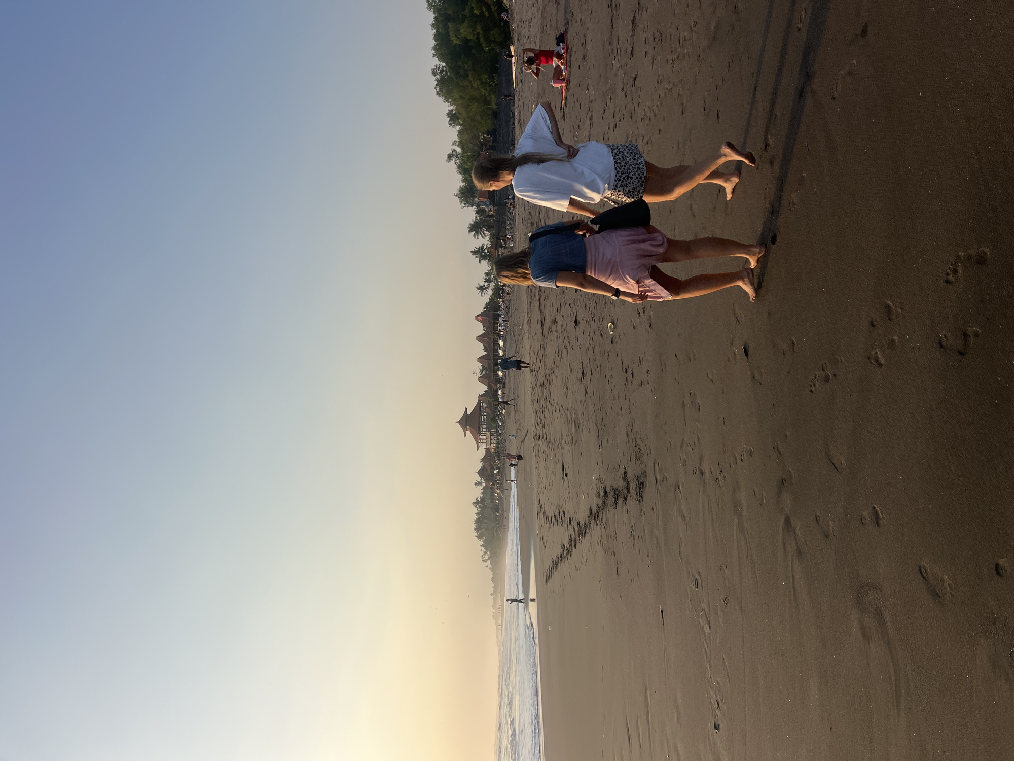 The image depicts a beachfront scene at sunset, likely in the city of Kediri, Indonesia, given the architectural style of the structures and vegetation visible. Two young women, viewed from behind, are walking barefoot on a dark sand beach towards the left of the frame. The woman on the left wears a blue t-shirt and a pink skirt, carrying a black shoulder bag. The woman on the right wears a white t-shirt and a leopard-print skirt.
In the background, several people are scattered across the beach, some near the water's edge and others further up the shore. A cluster of traditional Indonesian-style buildings with red-tiled roofs are visible along the treeline, suggesting a resort or tourist area. Gentle waves are breaking on the shore, and the sky exhibits a soft gradient of pale yellow and blue, indicating the sun is low on the horizon. Footprints are clearly visible in the wet sand near the water. To the right, a woman in a red top is seated on the sand, possibly with a pink object beside her. The overall atmosphere is serene and warm, characteristic of a tropical beach at dusk.