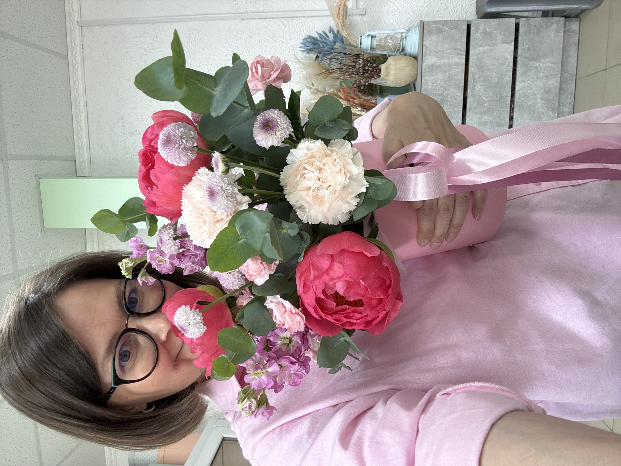 The image is a selfie of a woman with dark hair and glasses, holding a bouquet of pink and white flowers in front of her face. The flowers include peonies, chrysanthemums, carnations, and eucalyptus leaves. The bouquet is wrapped in pink paper and tied with a pink ribbon. The woman is wearing a pink shirt.
In the background, there is a wall with a textured surface and a green panel. There is also a gray crate and some dried flowers.
The location is in the city of Izhevsk, Russia.