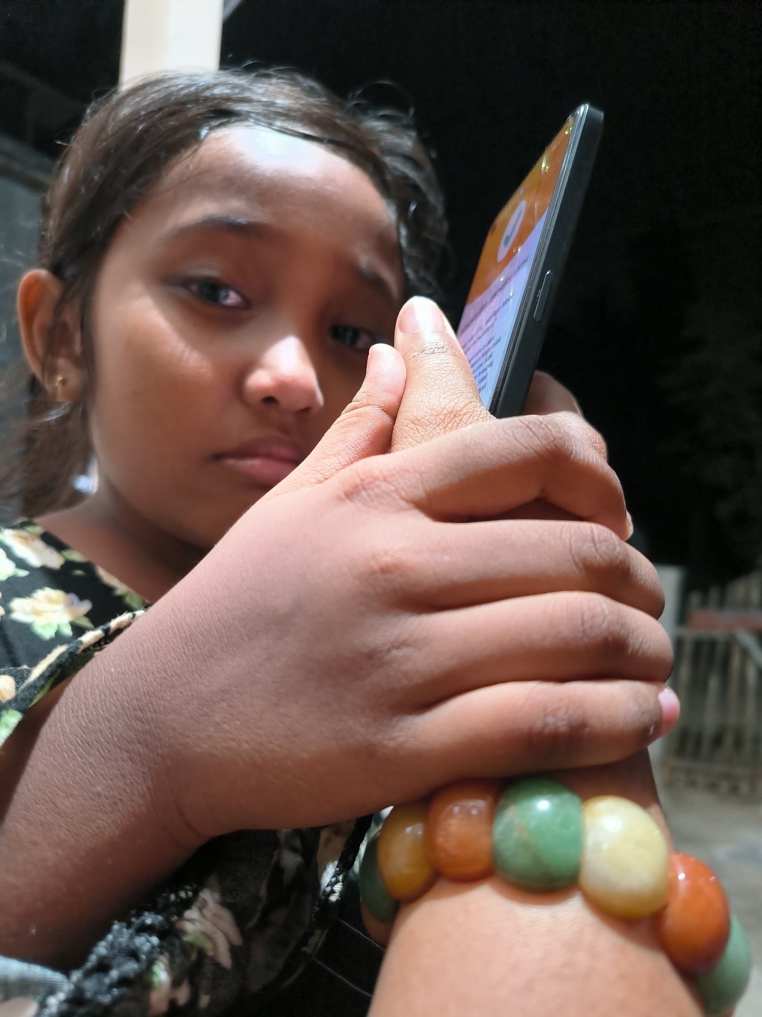 An outdoor, close-up shot features a young girl holding a smartphone in her hands. The girl, with dark hair and brown eyes, is looking down at the phone with a somber expression. Her right hand is holding the phone, while her left hand is wrapped around the phone and her right wrist. A colorful bracelet, made of several large, polished stones in shades of green, orange, and yellow, adorns her wrist. She is wearing a dark floral patterned shirt. The background is dark, suggesting it is nighttime. The lighting is artificial and casts shadows, highlighting the textures of her skin and the phone. The scene appears to be in a residential area, possibly a porch or yard, with hints of a fence or railing visible in the background. The image's setting is believed to be Lhokseumawe, Indonesia.
