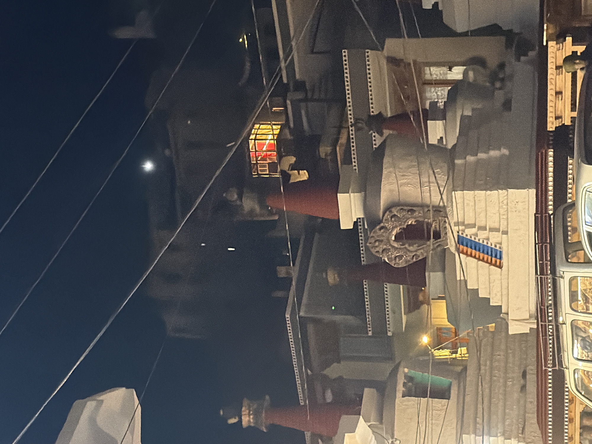 This image depicts a nighttime scene in Leh, India, likely near the Leh Palace or a similar religious structure. The foreground features the tops of several white, tiered stupas, characteristic of Tibetan Buddhist architecture. Some of these stupas are topped with red, conical spires. The structures have intricate decorative elements, including a carved archway on one stupa. In the background, buildings are visible, some with illuminated windows displaying warm yellow and red light, suggesting an interior light source. A faint moon is visible in the dark sky, along with several overhead wires. The lower portion of the image shows the roof rack of a white vehicle, and the heads of at least two individuals can be seen inside, suggesting the presence of onlookers or passersby. The overall atmosphere is serene and illuminated by artificial lights and the faint moonlight.
