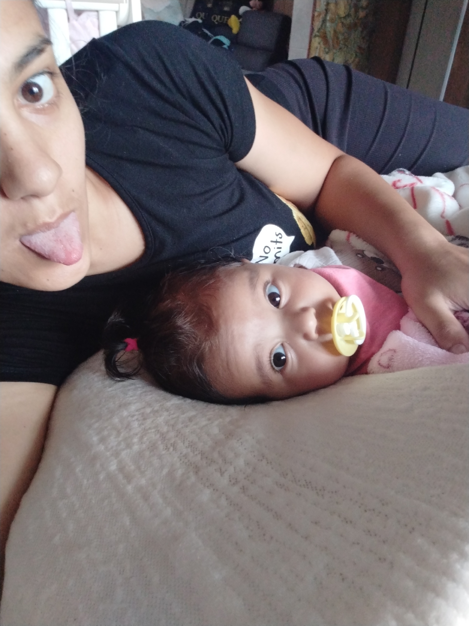 A selfie shows a woman with her tongue sticking out and a baby lying beside her. The woman is wearing a black t-shirt. The baby is wearing a pink outfit and has a yellow pacifier in its mouth. They are both lying on a bed with a light-colored blanket. The background is blurred but appears to be a room with furniture.