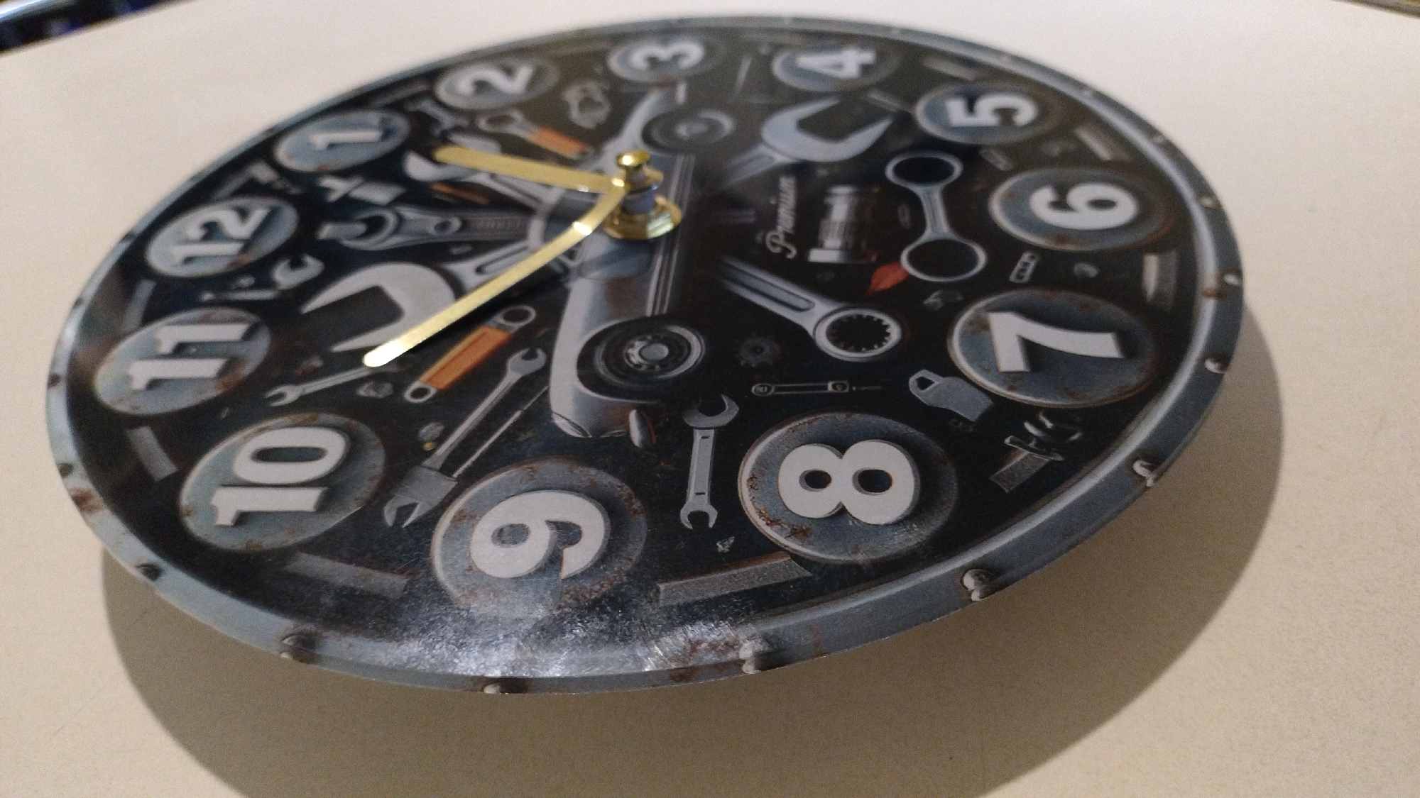 This media file shows a close-up, angled view of a round clock. The clock face is designed with a dark, distressed background and features images of various tools, such as wrenches and bolts, scattered across it. Large, white numerals represent the hours, with numbers 7 through 12 clearly visible. The numbers are enclosed in oval shapes with a distressed, metallic appearance. The clock has two hands: a thinner, longer gold-colored hand pointing approximately to the 1 o'clock position, and a shorter, thicker gold-colored hand pointing slightly past the 10 o'clock position. The clock has a thick, gray bezel with visible rivets around the edge. There is some visible text on the clock face that appears to say "Premium." The lighting suggests an indoor setting, possibly with artificial light, creating a shadow on the right side of the clock. The overall aesthetic of the clock suggests a theme related to automotive mechanics or a workshop.