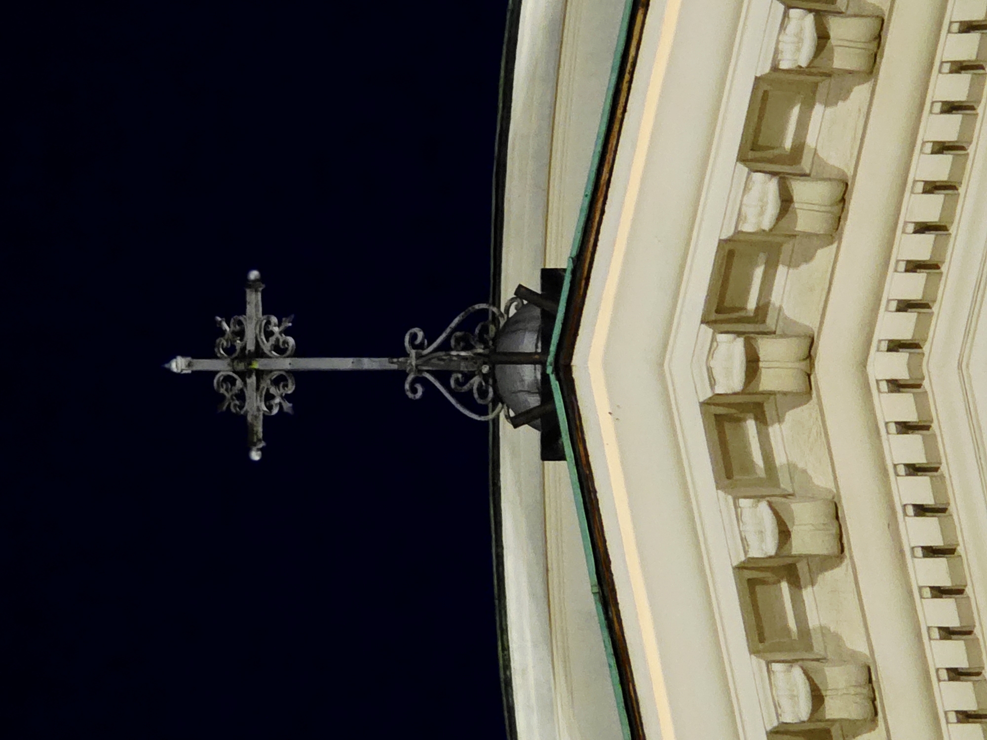 The image shows the upper portion of a building, likely a church, at night. A decorative metal cross with ornate flourishes is positioned centrally at the apex of the building's facade. The cross is silhouetted against a dark night sky. Below the cross, architectural details of the building are visible, including a curved roofline, decorative molding, and what appear to be corbels or brackets supporting the roof. The building's exterior is a light color, possibly stone or painted plaster. The overall impression is one of solemnity and architectural grandeur. This building is the Church of the Holy Savior in Warsaw, Poland.