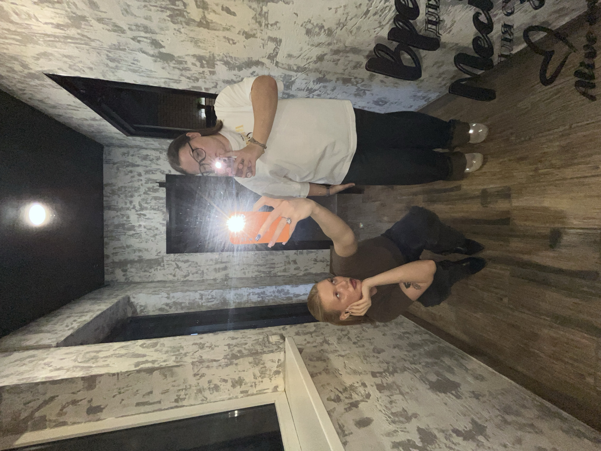Two people are taking selfies in what appears to be a room with unconventional architecture. The room has light-colored, textured walls and dark wood-like flooring. One person, wearing a white shirt and dark pants, is standing and taking a selfie in a mirror or reflective surface. This person wears glasses. The second person is lying down, holding up a phone to take a selfie. This person is wearing a brown shirt and dark pants. The room contains windows of varying shapes. The image includes a partially visible decorative sign in Russian. The location is Chaikovsky, Russia.