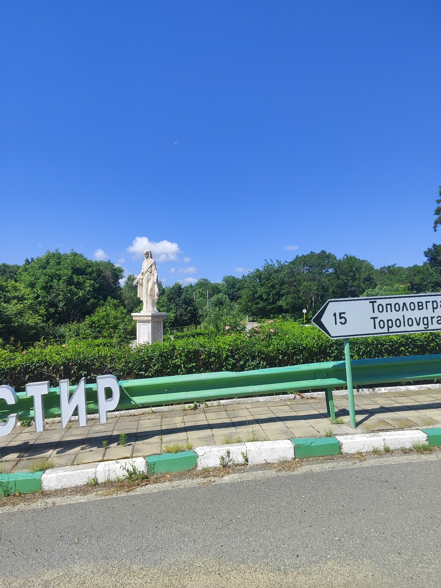 The image depicts a bright, clear day with a vibrant blue sky and a crescent moon visible. In the foreground, a green metal guardrail runs horizontally across the frame. To the left, large white three-dimensional letters spell out "СТИР," partially obscured by the guardrail. The ground beneath the letters is paved with square concrete tiles, bordered by a curb painted with alternating green and white sections.
Behind the guardrail and letters, a manicured hedge forms a dense green backdrop. To the left, a white statue of a robed figure stands on a pedestal amidst the foliage. The statue appears to be a historical or religious monument. A directional signpost is visible to the right, indicating "15" and "Тополовгра Тороlovгра" with an arrow pointing to the right, suggesting a distance and a destination likely within or near Golyam Manastir, Bulgaria. The background is filled with lush green trees, indicative of a park or natural setting. The overall scene suggests a roadside view in a scenic location.