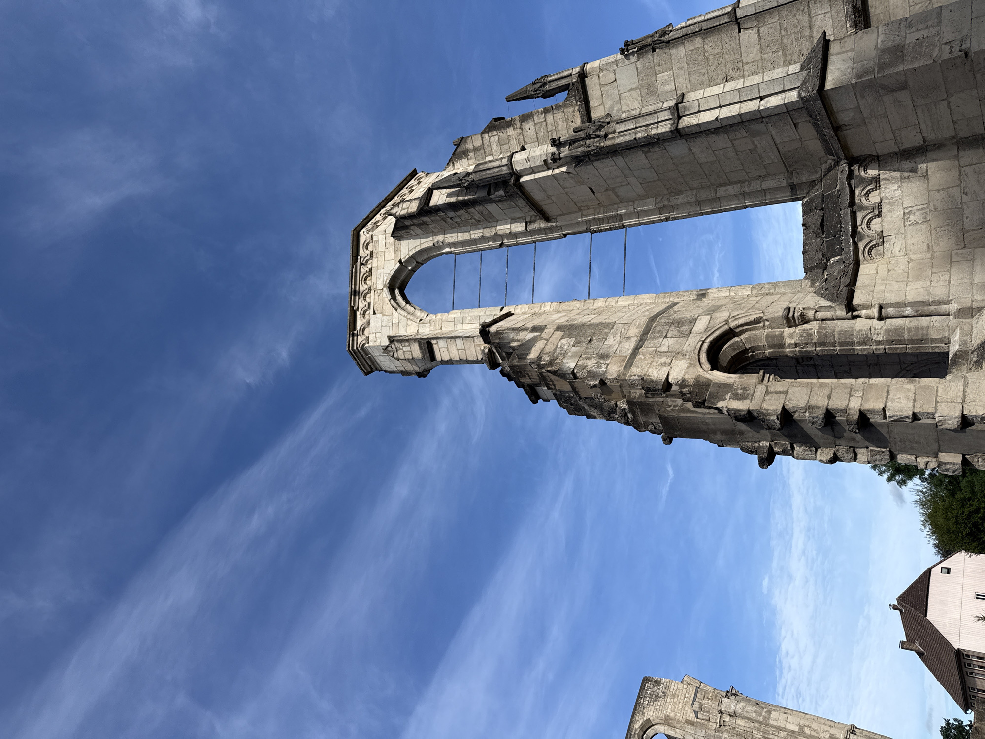 In Walkenried, Germany, the skeletal remains of a Gothic structure stand against a bright blue sky dotted with thin, wispy clouds. The ruins are constructed of weathered grey stone, with grand arched openings framing the sky. The most prominent feature is a tall, angular section of the building, its stone blocks showing the wear of time. This section has a large, pointed archway at its apex, with horizontal metal bars spanning the void within. To the left, a smaller, curved archway is visible in another section of the ruins. In the lower left of the frame, a portion of a light pink building with a dark roof peeks out from behind the ruins. Green trees are visible in the mid-ground, nestled between the modern building and the ancient stone. The perspective is from a low angle, looking up towards the sky, emphasizing the height and grandeur of the ruined architecture. There are no people visible in the scene.
