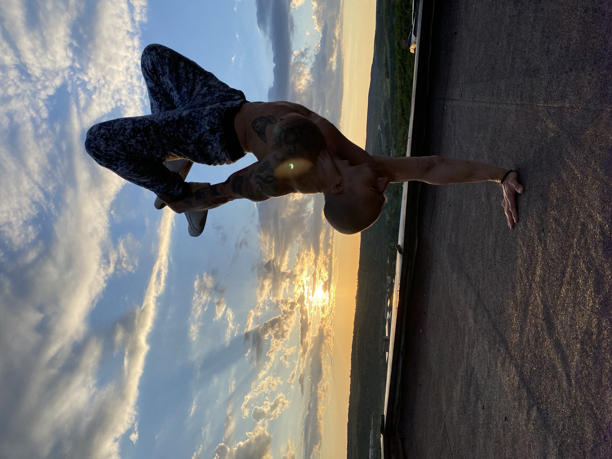 A shirtless man in mottled pants is performing a handstand on what appears to be a rooftop at sunset. He is leaning forward, his outstretched hand on the ground, while his legs are bent and raised in the air. The man has tattoos on his arms and torso, and his head is shaved. The setting sun is visible in the background, along with clouds and a view of a city. The rooftop is textured and dark. The location is Pleven, Bulgaria.