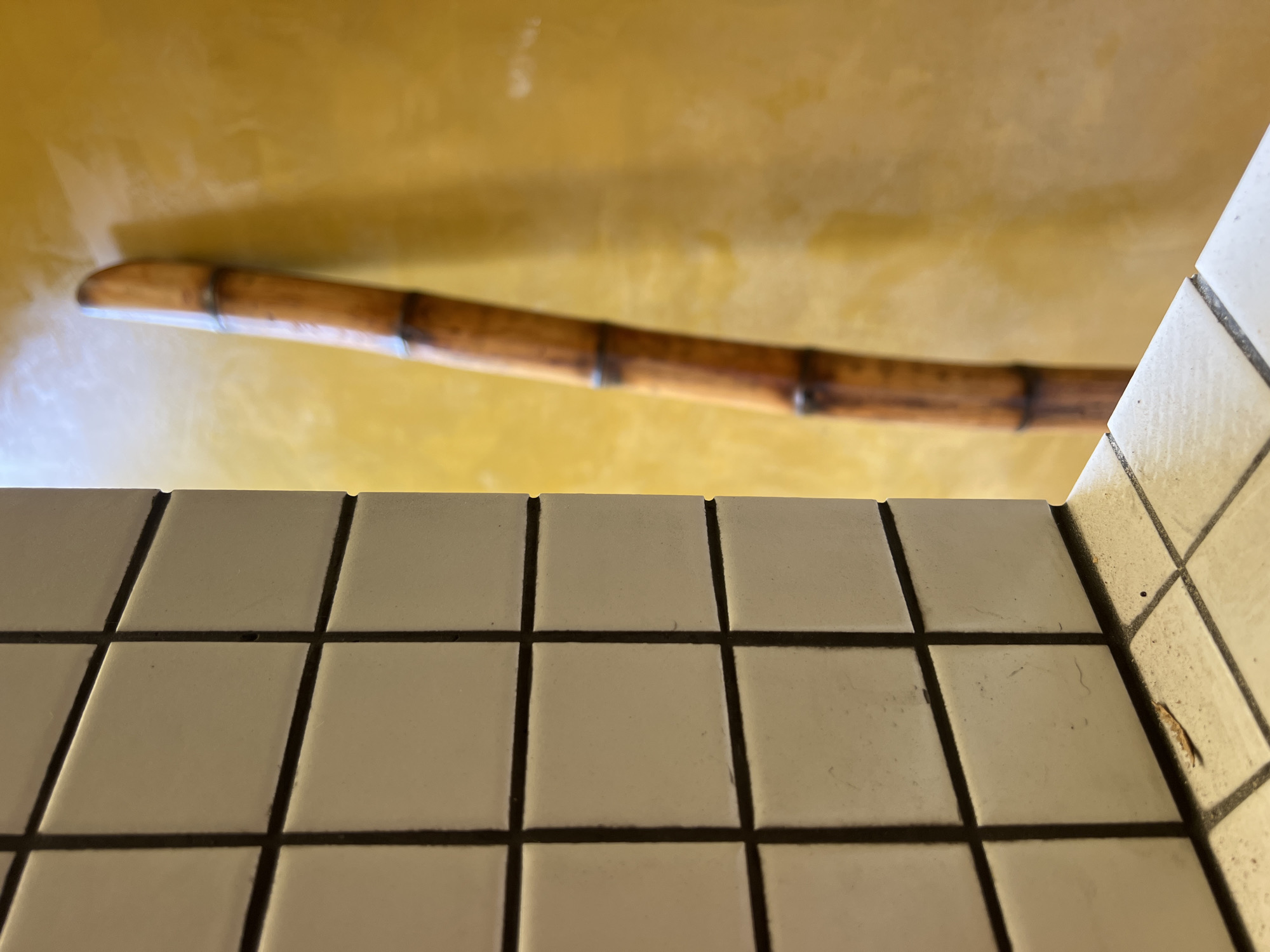 The image shows a close-up, eye-level view of a tiled corner of a room. The tiles are small, square, and cream-colored with dark grout. On the left side of the image, the tiled wall recedes into the background, revealing another tiled surface at a right angle. To the right of the tiled corner, a thin, brown, bamboo-like object, possibly a walking stick or staff, is leaning against a textured, yellow wall. The bamboo object has dark bands at intervals along its length. The lighting suggests the scene is indoors, possibly in a hallway or a room with a warm ambiance.