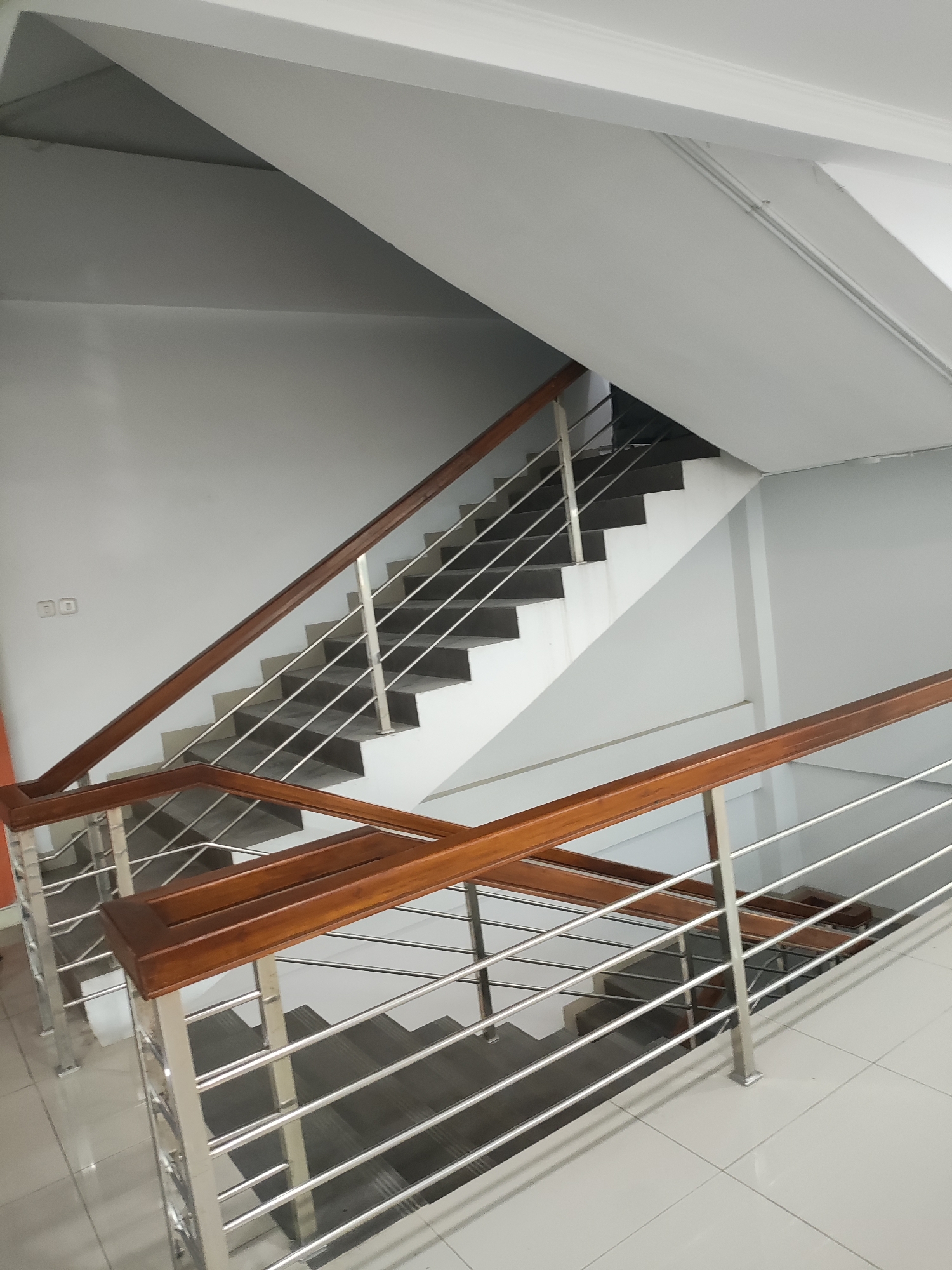 This image shows an indoor staircase with wooden handrails and stainless steel railings. The walls are painted white, and the floor is tiled with white square tiles. The staircase appears to be part of a modern building, possibly an office or residential building. There are no people visible in the image. The location is specified as Cirebon, Indonesia.