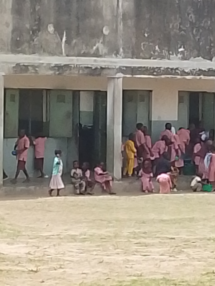 The image depicts a crowded outdoor scene in Igbo Ijaiye, Nigeria, showing numerous children gathered in front of a two-story concrete building. The building's upper facade is weathered and discolored, while the lower level features multiple rectangular doorways or windows with dark, possibly green, panels. A raised concrete platform or step runs along the base of the building, serving as a seating area for some individuals. The ground in the foreground is an unpaved, light-colored, dry dirt or dusty area, with minimal visible vegetation.
Approximately twenty-five to thirty individuals, predominantly children aged roughly 5-12, are visible. Most children are dressed in similar light-red, pink, or maroon-colored short-sleeved shirts and shorts or skirts, consistent with school uniforms. Notable exceptions include one child on the far right wearing a yellow long-sleeved outfit and another child in the mid-left foreground wearing a light blue/green top and a cream-colored skirt. One boy seated on the left wears a camouflage-patterned top and shorts. An adult-like figure or older child in a dark blue/black shirt is seen from the back among a cluster of children on the right.
Actions include children standing, walking, and sitting. On the far left, two boys in pink/maroon uniforms are standing, with one appearing to walk towards the left. In the mid-left, a small group of children, including the one in the light blue/green and cream outfit, are seated on the concrete platform. On the right side, a larger group of children is densely clustered around an entrance, some appearing to be in motion or interacting, suggesting a period of recess or transition between classes. No specific objects beyond clothing are clearly identifiable due to the low resolution of the image. The scene is illuminated by natural daylight, casting no distinct shadows.