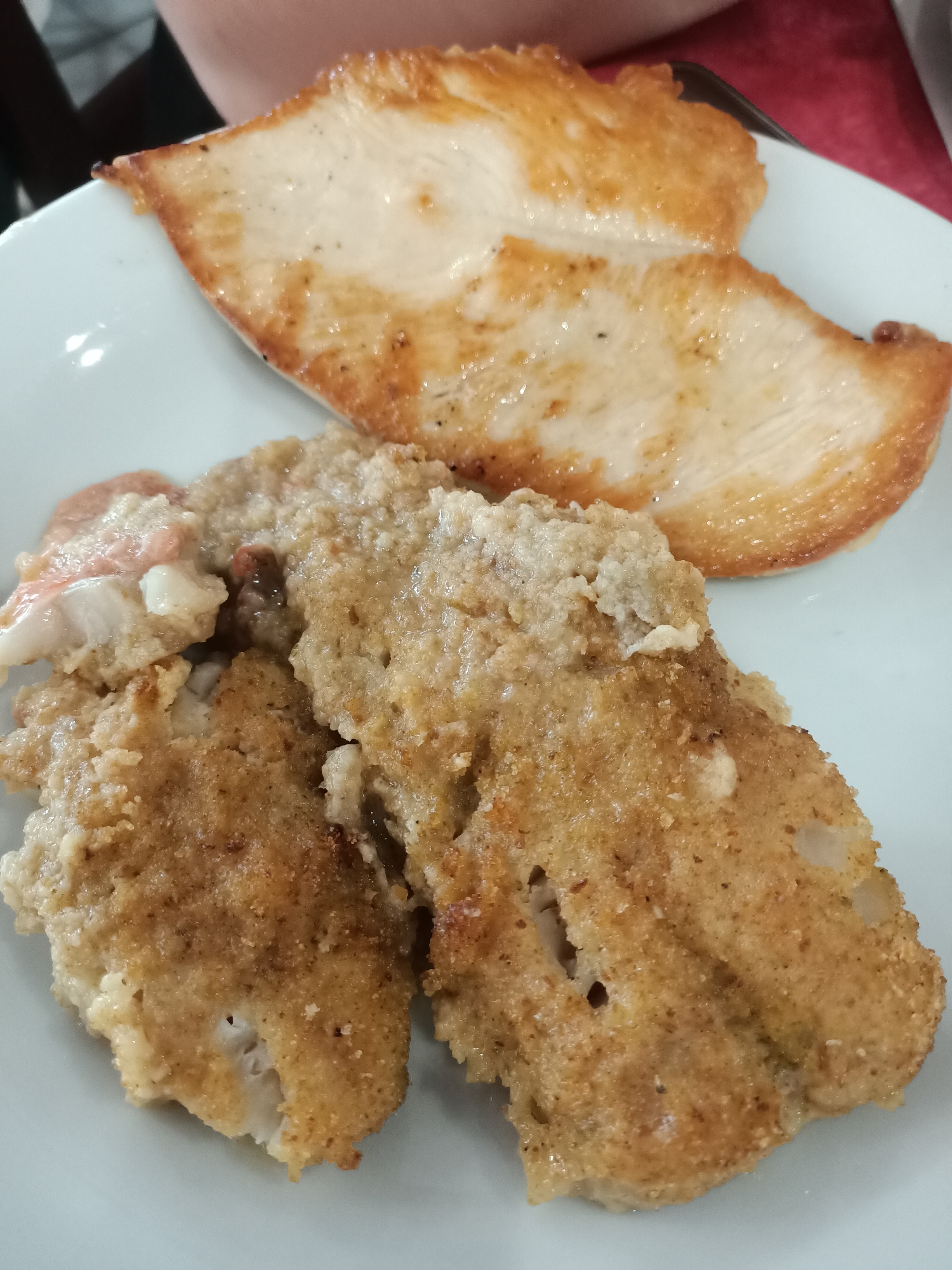 A white plate holds two servings of food. In the upper portion of the plate, two thin slices of cooked white meat, possibly chicken or turkey breast, are arranged overlapping each other. The meat is lightly browned and appears to have been pan-fried or grilled. Below these slices are two larger, thicker pieces of breaded and fried food, likely fish fillets or chicken tenders. These pieces have a golden-brown, crispy crust. A person's arms and shoulders are visible in the upper left and upper right corners of the image, suggesting they are seated at a table and about to eat. The setting appears to be a restaurant or dining area.