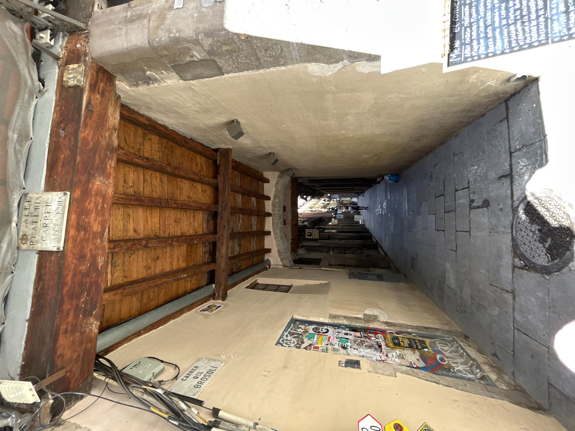 The image depicts a narrow, cobblestone alleyway in Barcelona, Spain, looking towards a street lined with buildings and lamps. Wooden beams support a covered section of the alley overhead. The walls of the alley are plastered and weathered, with one side covered in a collage of stickers and graffiti. A street sign reads "Carrer del Brotsoli". Further down the alley, a few individuals are visible, including a person walking away from the camera and another on a bicycle. On the right side of the alley, a chalkboard menu is propped up, advertising food items in Spanish. The overall impression is of a historic and bustling urban environment.