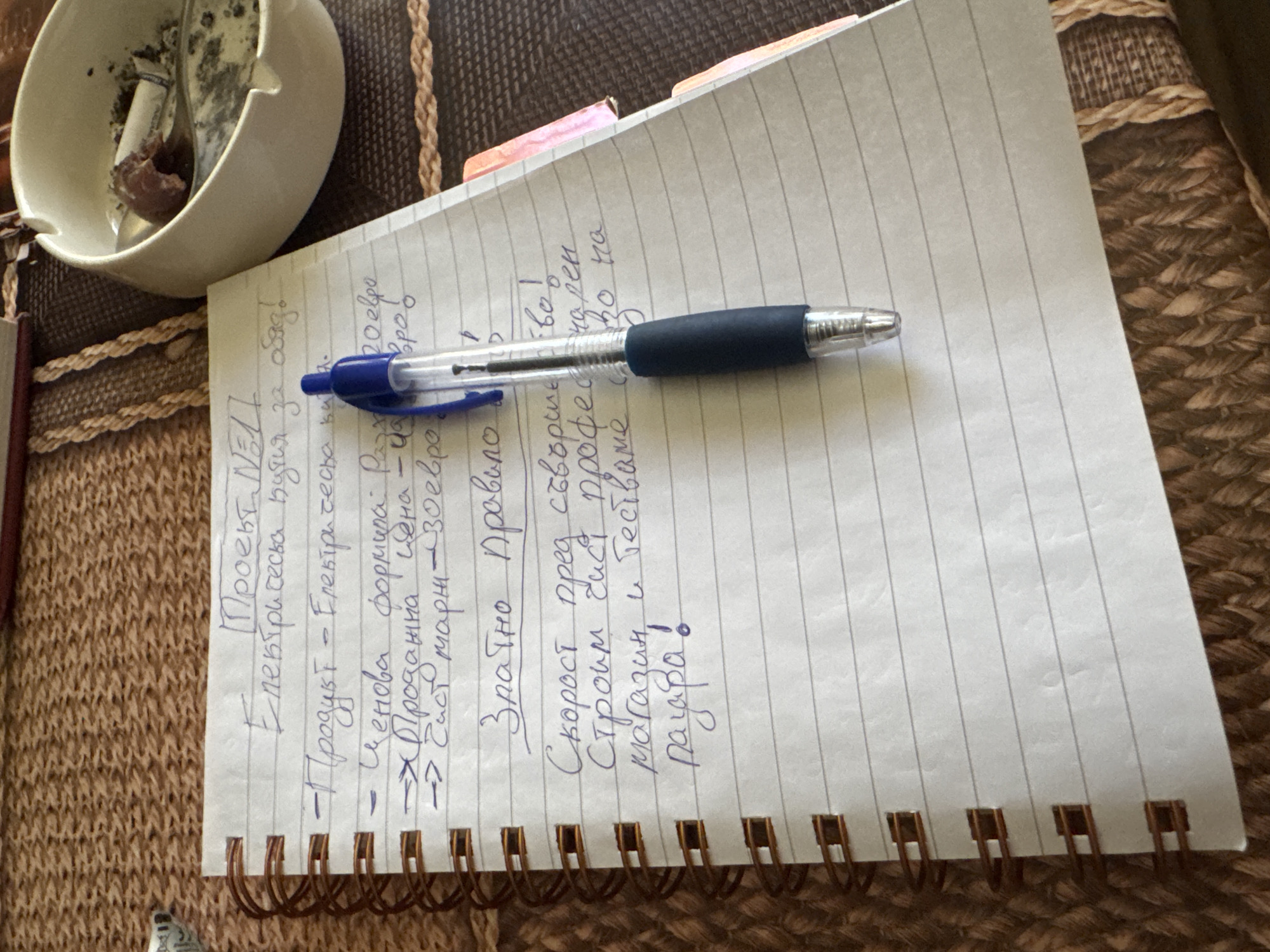 The image shows a close-up, eye-level view of a lined notepad on a textured brown surface. A blue and silver pen rests on the notepad, pointing downwards. The notepad contains handwritten text in blue ink, which appears to be a list of tasks or points related to a project. The text is partially legible but includes phrases like "Проект №11," "Електрическа индустрия за обяд," "Продукт – Електрическа," "Ценова формула," "Продажна цена," "Чист марж," "30 евро," "Здратно Правило," "Скорост," "Строим," "магазин," "тестваме," and "научава." A white ceramic ashtray with cigarette butts and ash is visible in the upper right corner of the image, suggesting a smoking area or a break. The background texture resembles a woven placemat or table runner. The scene does not provide any specific information about the city of Sofia, Bulgaria, or any identifiable landmarks.