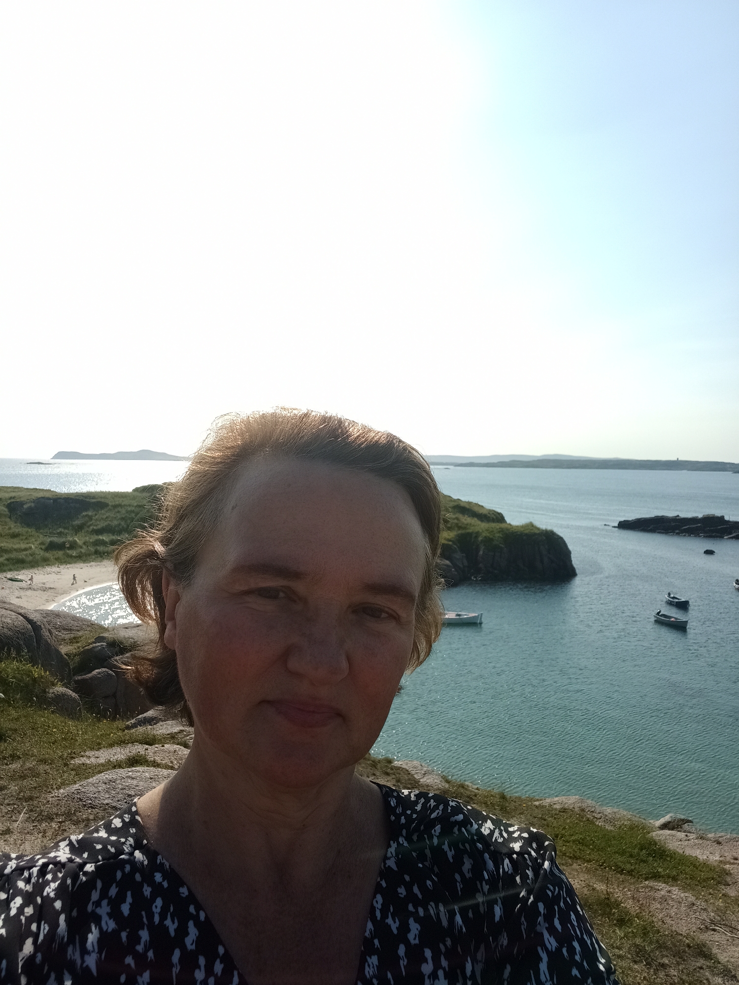 A woman takes a selfie from a rocky, grassy incline overlooking a bay in Carnboy, Ireland. The woman, facing forward, has light brown hair pulled back and is wearing a black and white patterned dress. The bay, which appears calm, contains several small boats anchored in the turquoise water. In the distance, a sandy beach can be seen with a few figures walking. Islands and hills form the horizon under a bright, clear sky.