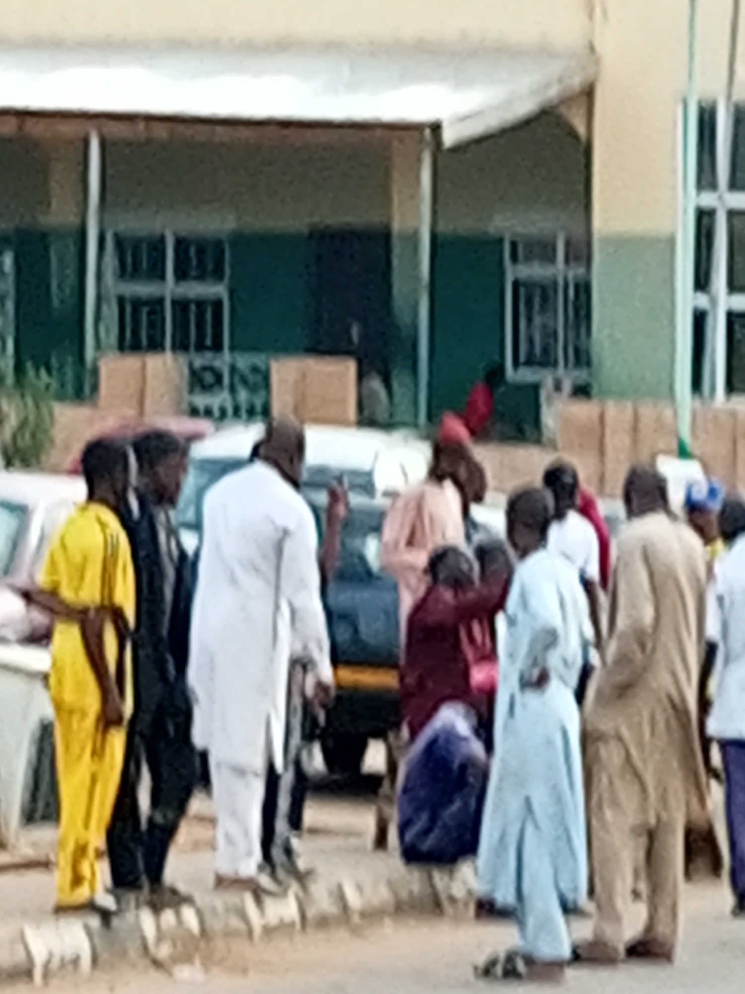 In Gombe, Nigeria, a group of people stand around what appears to be vehicles on a paved surface. The setting seems to be outdoors, possibly near a building with windows and a covered walkway. The individuals are dressed in a variety of clothing, with some wearing traditional attire like flowing robes and head coverings, while others are in more casual wear. One person in the foreground wears a bright yellow outfit, while another is in a white robe. The group appears to be engaged in conversation or observing something. The lighting suggests it could be daytime, possibly late morning or afternoon. The overall atmosphere seems neutral, with no strong indicators of specific emotions or a particular event. There is no visible text in the image.
