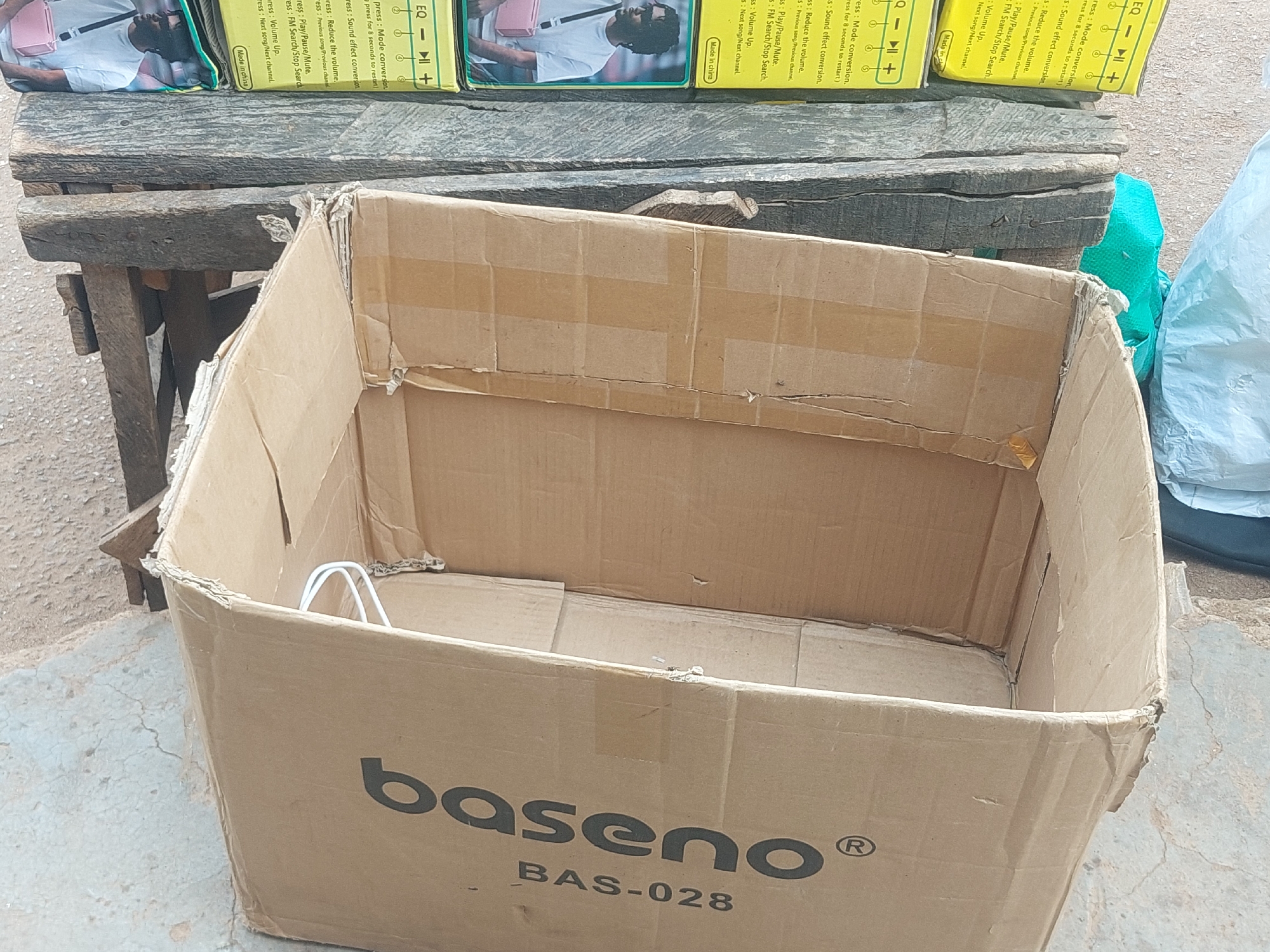 A cardboard box is positioned in the foreground, its sides partially collapsed. The box is a light brown color and features black text that reads "baseno" and "BAS-028". A white wire loop is visible inside the box. In the background, a wooden structure with multiple planks is visible, upon which several items with yellow packaging and images of people are stacked. The ground is composed of cracked concrete. The environment appears to be outdoors.