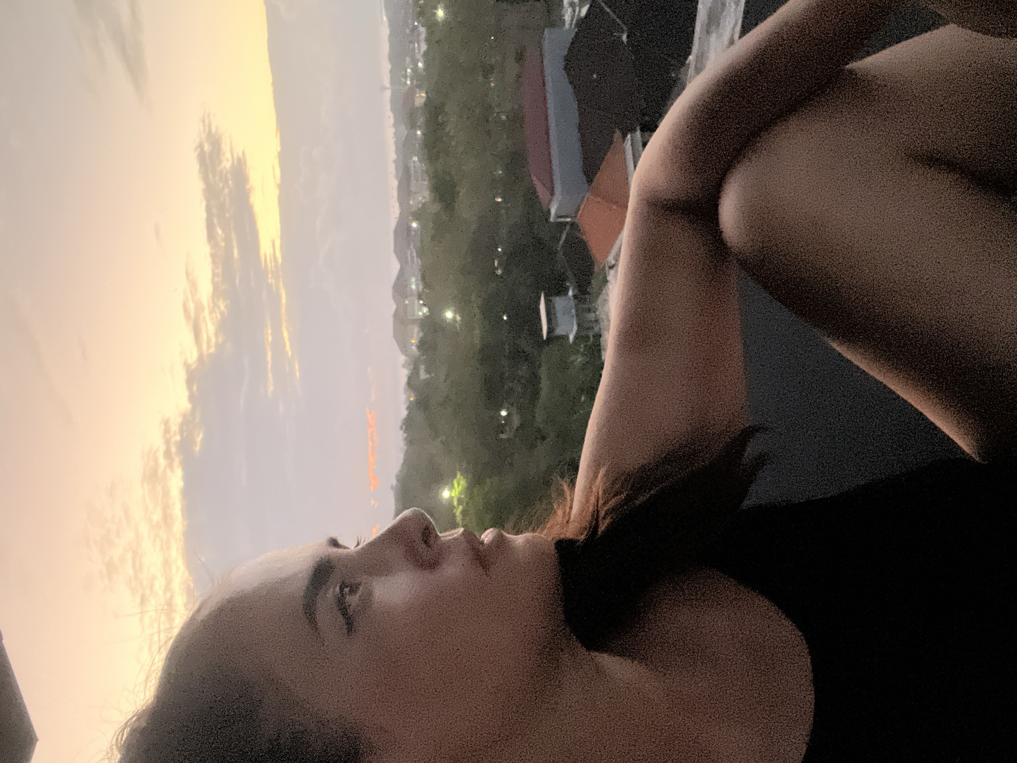 A woman is captured in a close-up profile shot, her head turned to the left, gazing into the distance. She has dark hair pulled back, and is wearing a black top. Her arm is bent, resting on her leg, and her knee is visible. The background depicts a hazy cityscape under a soft, pastel-colored sky, suggesting either dawn or dusk. The city appears to be Kuta Selatan, Indonesia, with visible rooftops, trees, and scattered lights. The overall mood is serene and contemplative.