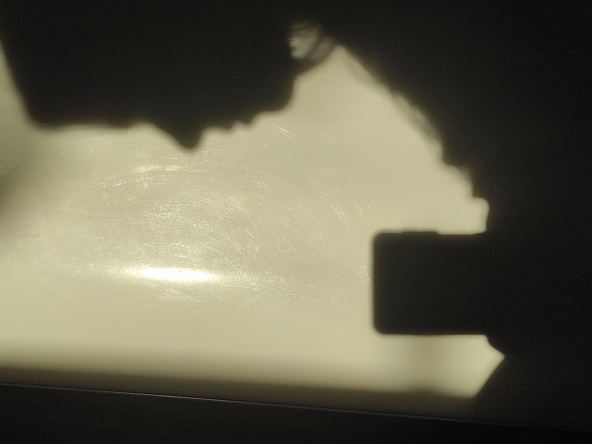 The image shows a shadow of a person taking a picture with a smartphone. The shadow is cast on a light-colored surface, possibly a table or counter. The person's head and shoulder are visible in the shadow, along with the outline of the phone. There is a bright spot of light reflected on the surface. The bottom of the image contains a dark horizontal line, possibly indicating the edge of the surface. The location is unknown.