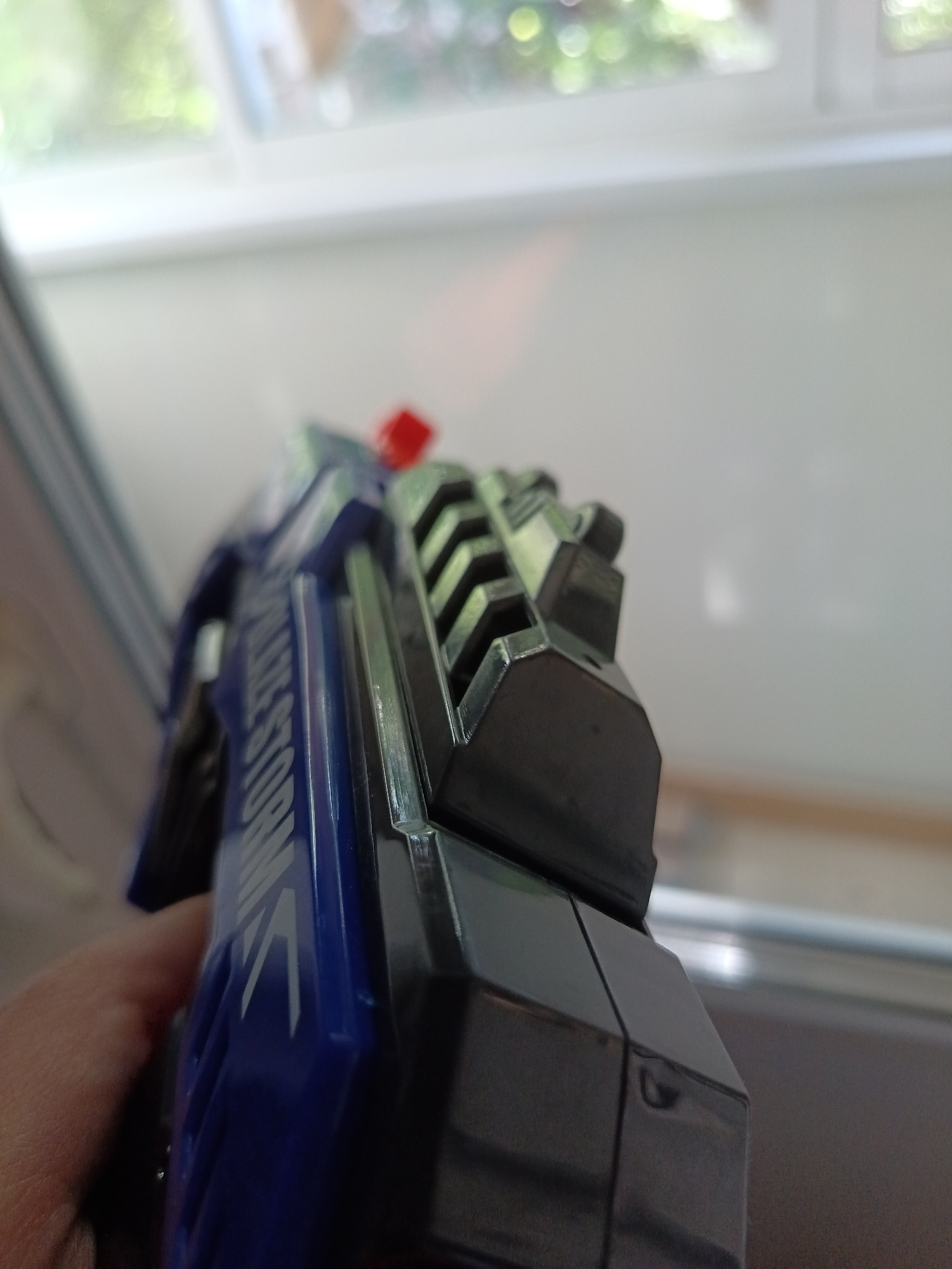 The image shows a close-up of a blue and black toy gun, possibly a Nerf gun, held in a person's hand. The gun has text printed on its side which appears to read "STORM". The background is blurred and appears to be an indoor setting with a window visible, showing greenery outside. There are no discernible landmarks or elements that would indicate a specific location in Minsk, Belarus. The lighting suggests it is daytime. The scene is static and there is no action taking place.