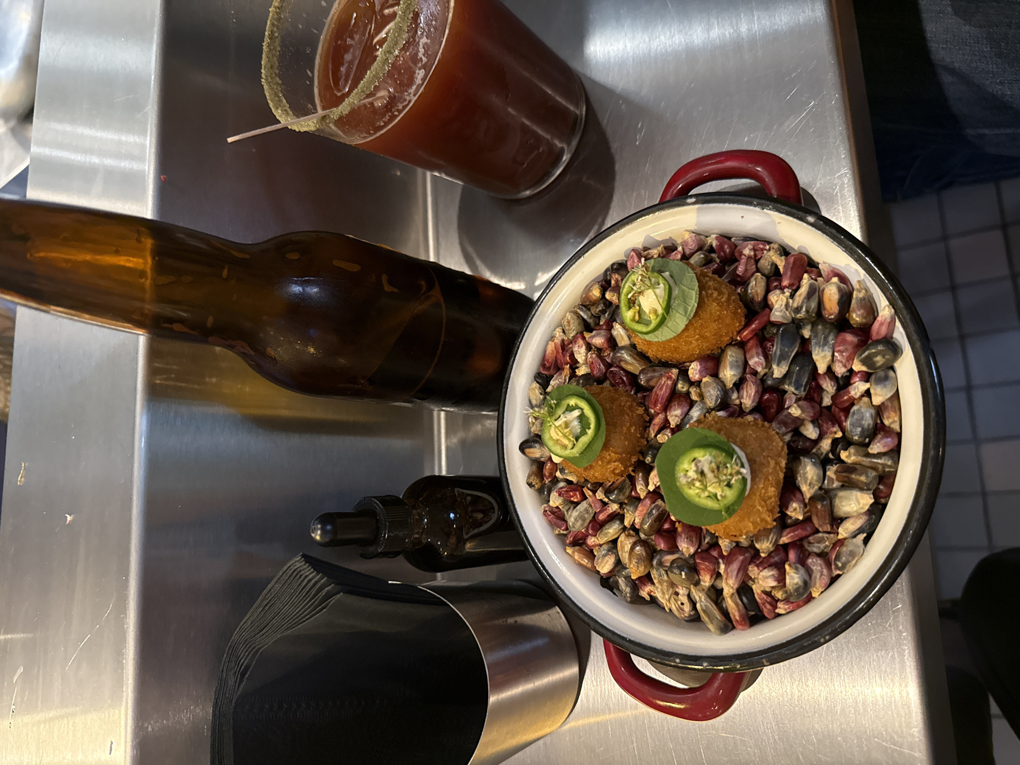 The image shows a close-up, overhead shot of food and drinks, possibly at a restaurant or bar in Mexico City, Mexico. The scene includes a tall glass of dark red liquid, likely a beer cocktail, with a rim coated in what appears to be salt. Next to it is a dark brown beer bottle, laying horizontally. There is also a small brown bottle with a dropper. The main focus is a white and red enamel pot filled with multi-colored kernels of corn and three fried food items. The food items are topped with a slice of jalapeño pepper and a small amount of white filling. The image also shows a stack of dark-colored napkins in a metal container. The background suggests a metal countertop or surface.