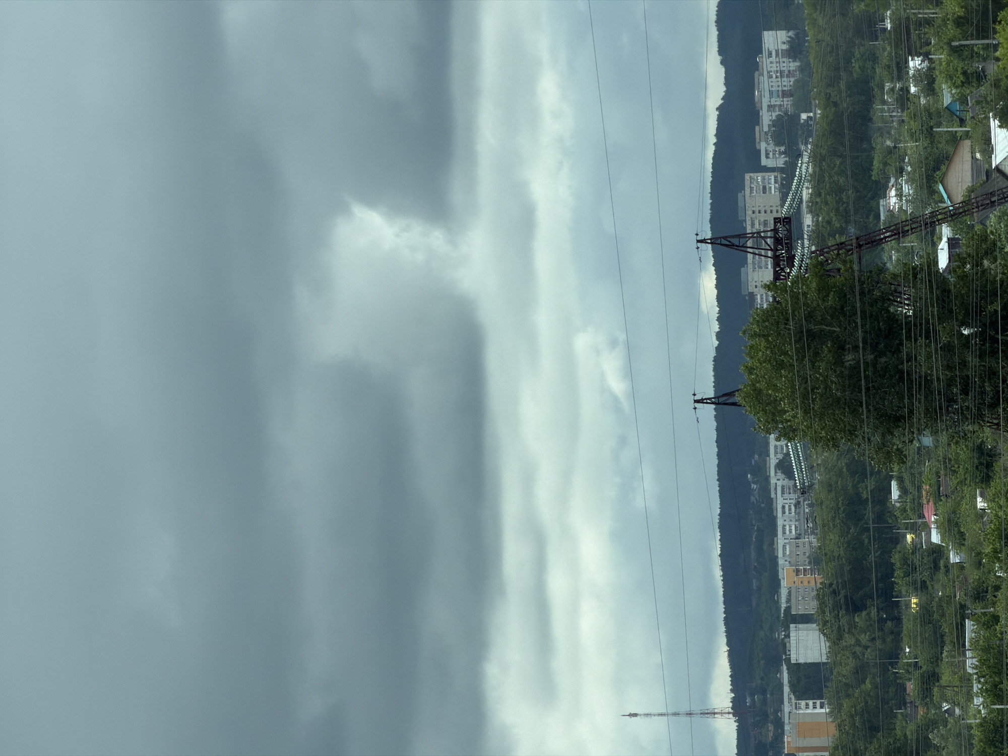 The image presents an expansive view of an urban or suburban landscape in Chaikovsky, Russia, under an overcast sky. The upper half of the frame is dominated by a dense, grey cloud cover, featuring areas of lighter grey and a prominent, irregular, vertically elongated bright cloud formation in the upper right quadrant.
In the foreground, a dense canopy of green trees and foliage occupies the lower portion of the image, partially obscuring residential structures. Multiple dark, horizontal power lines traverse the scene at various elevations. On the right side, a substantial section of a dark red or maroon lattice electrical transmission tower is visible, extending upwards into the midground.
The midground reveals several multi-story residential apartment buildings, predominantly light grey or white, with one structure featuring prominent yellow sections. These buildings are situated amidst further green trees and shrubbery, appearing to be built on rolling hills.
The background is defined by a dark green, densely forested mountain or hill range stretching across the horizon. A tall, slender communication tower, distinguished by red and white striped sections, rises prominently from the left side of the distant landscape. No individuals are visible in the scene.
